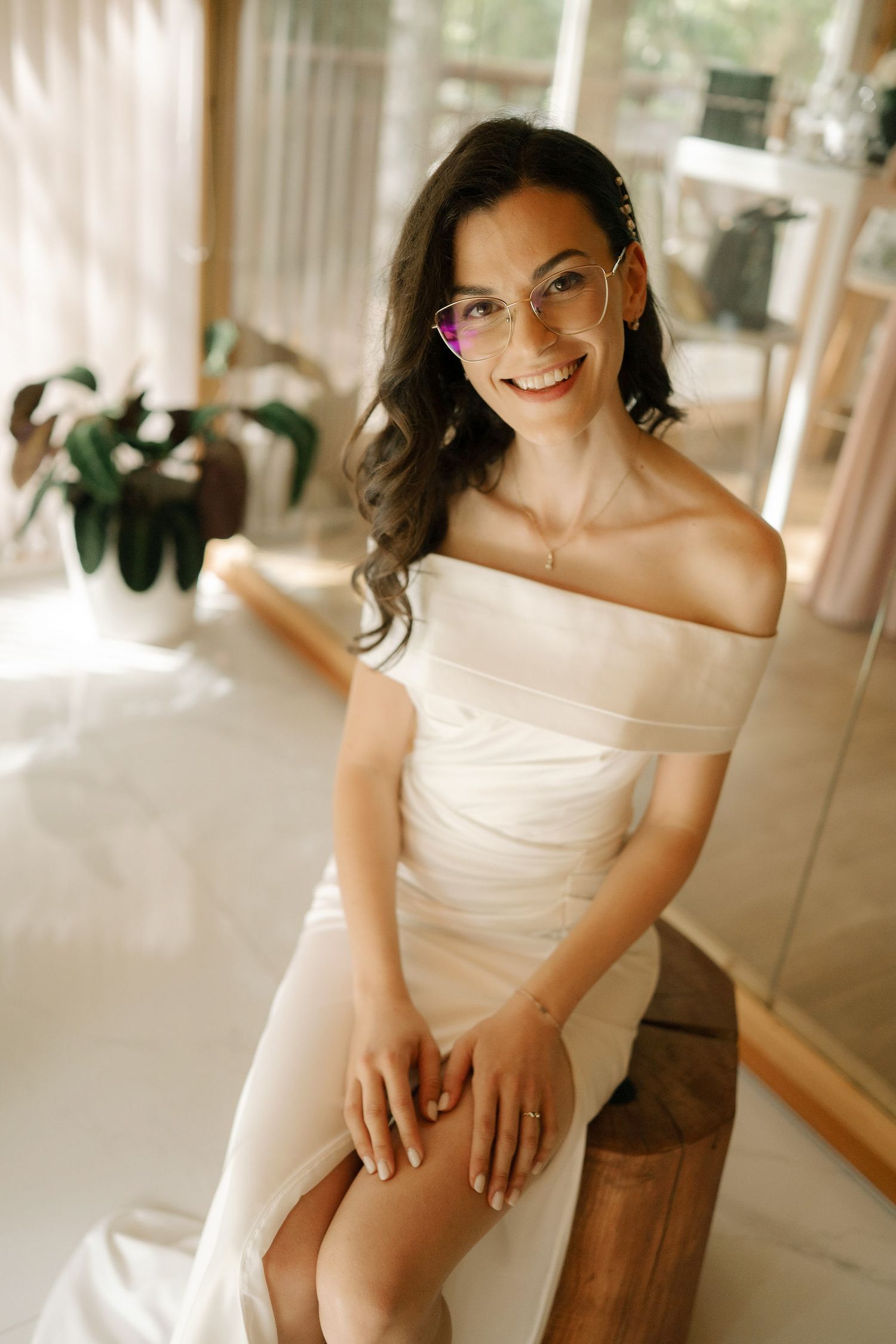A bride in an off-shoulder white wedding dress sits elegantly on a wooden stool, wearing glasses and smiling warmly.