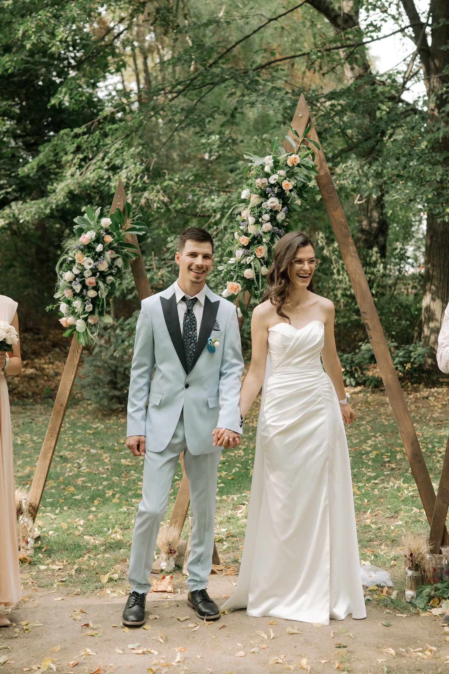A wedding couple walks together outdoors along a path with trees and scattered autumn leaves.