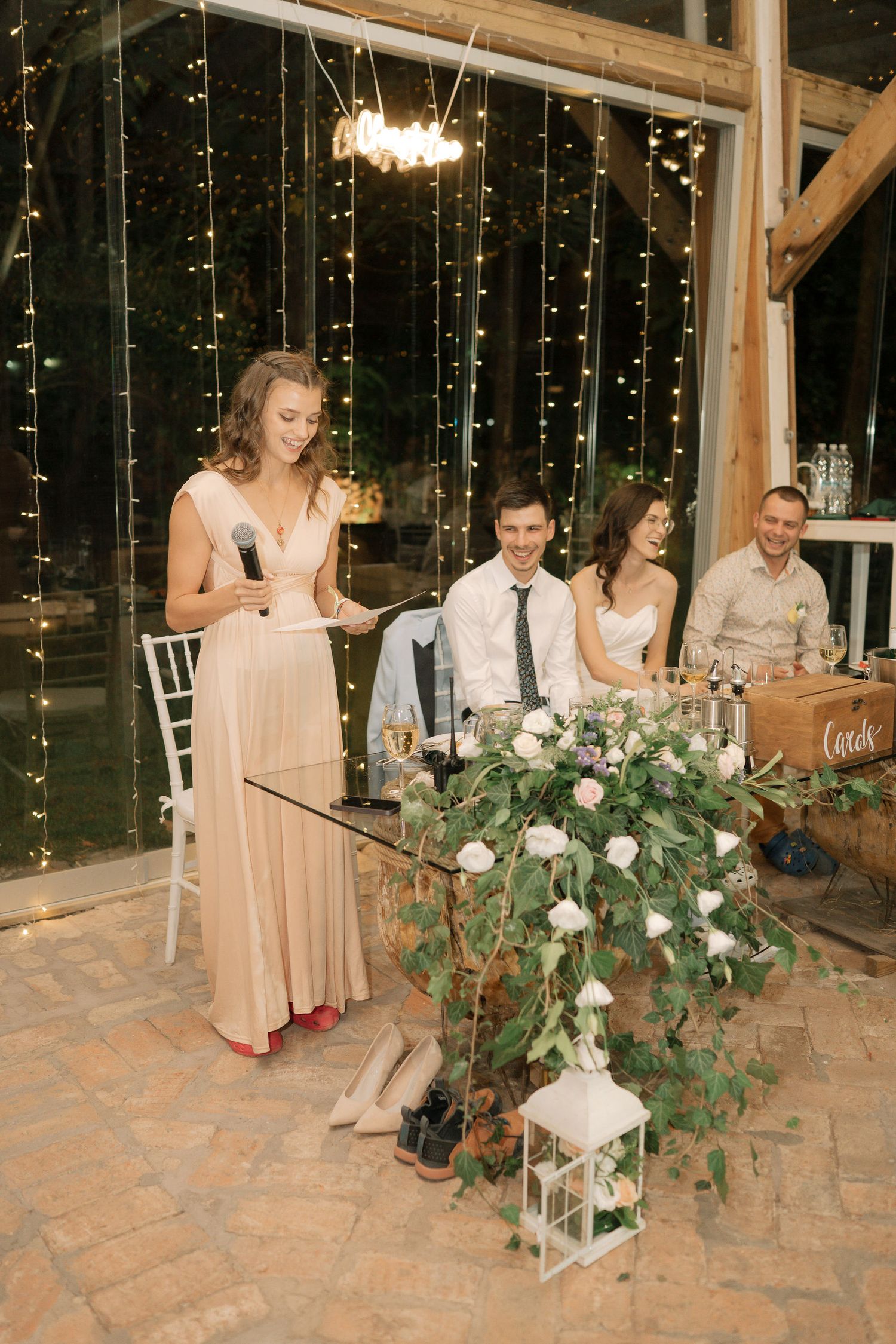 Wedding reception guests gathered around elegant table settings with floral centerpieces and lantern decorations at evening venue.