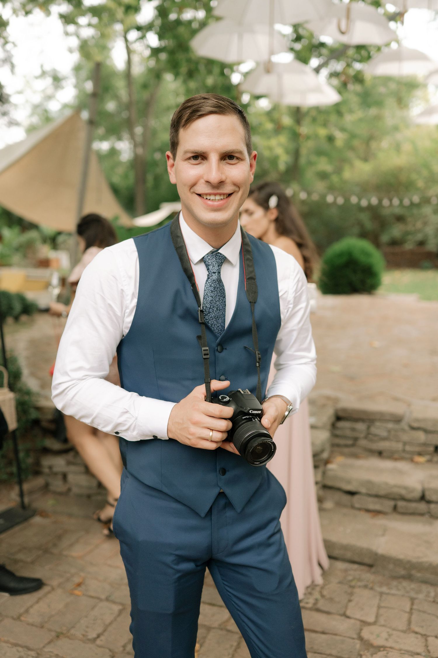 Wedding photographer in navy vest and tie captures moments on camera during outdoor reception.