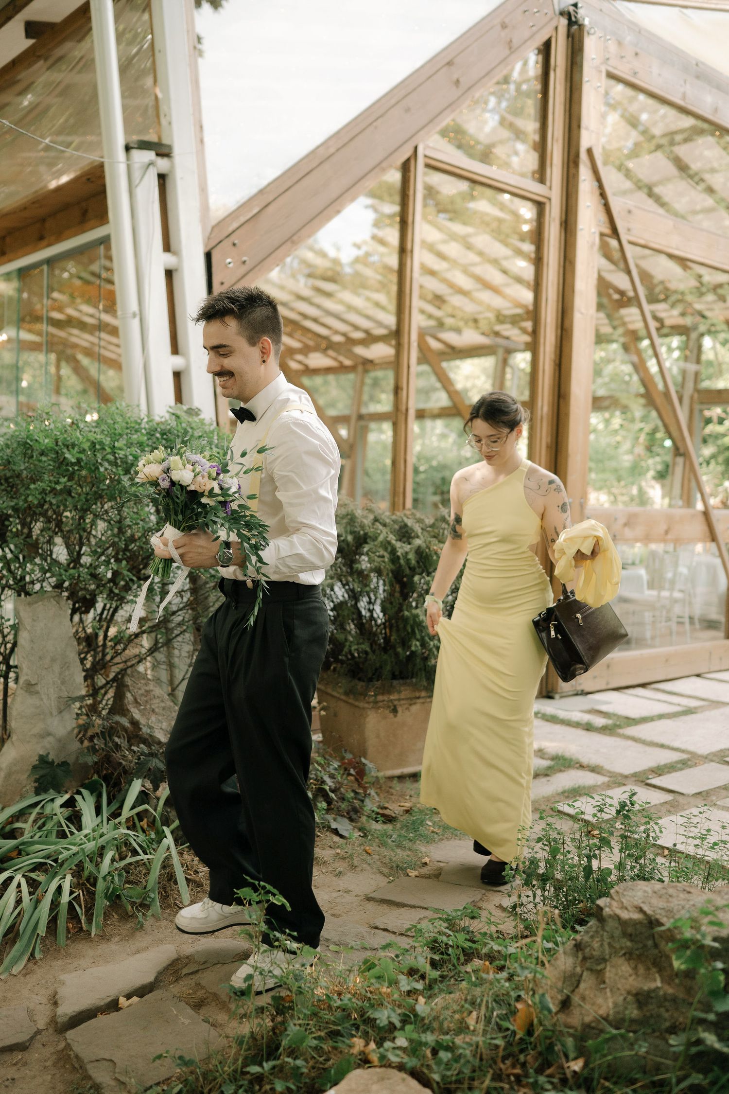 Two people stand in a sunlit greenhouse surrounded by plants and greenery in a casual outdoor setting.