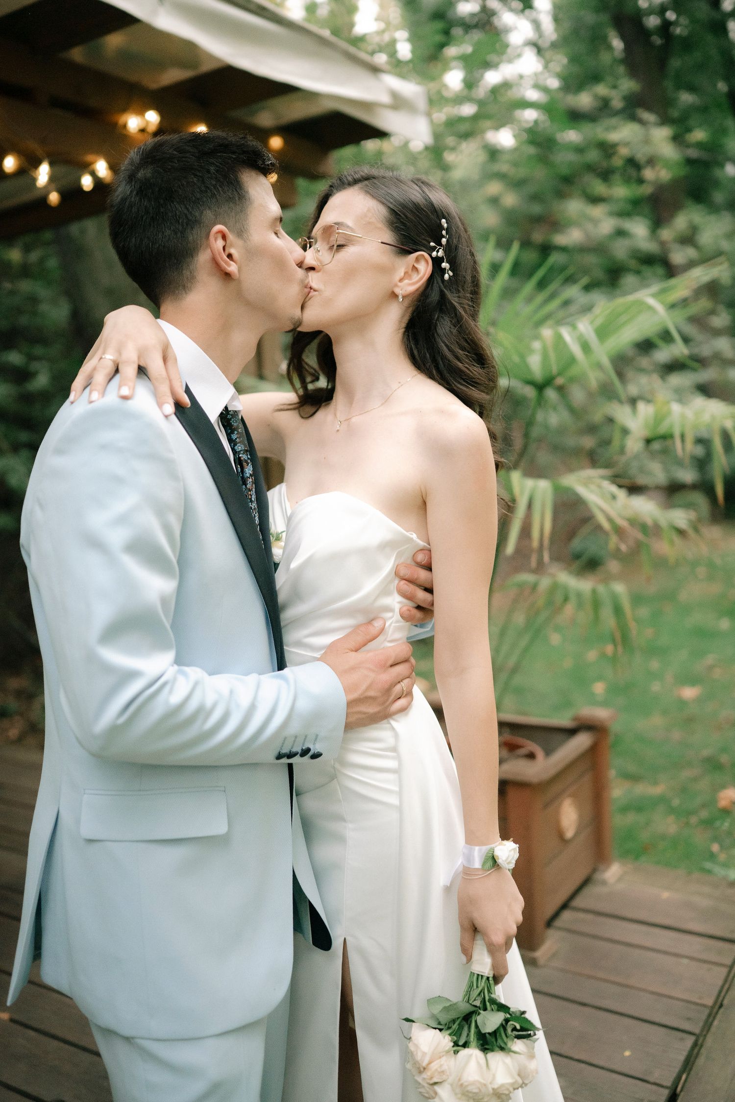 A romantic wedding couple shares intimate kisses on a wooden deck surrounded by lush greenery.