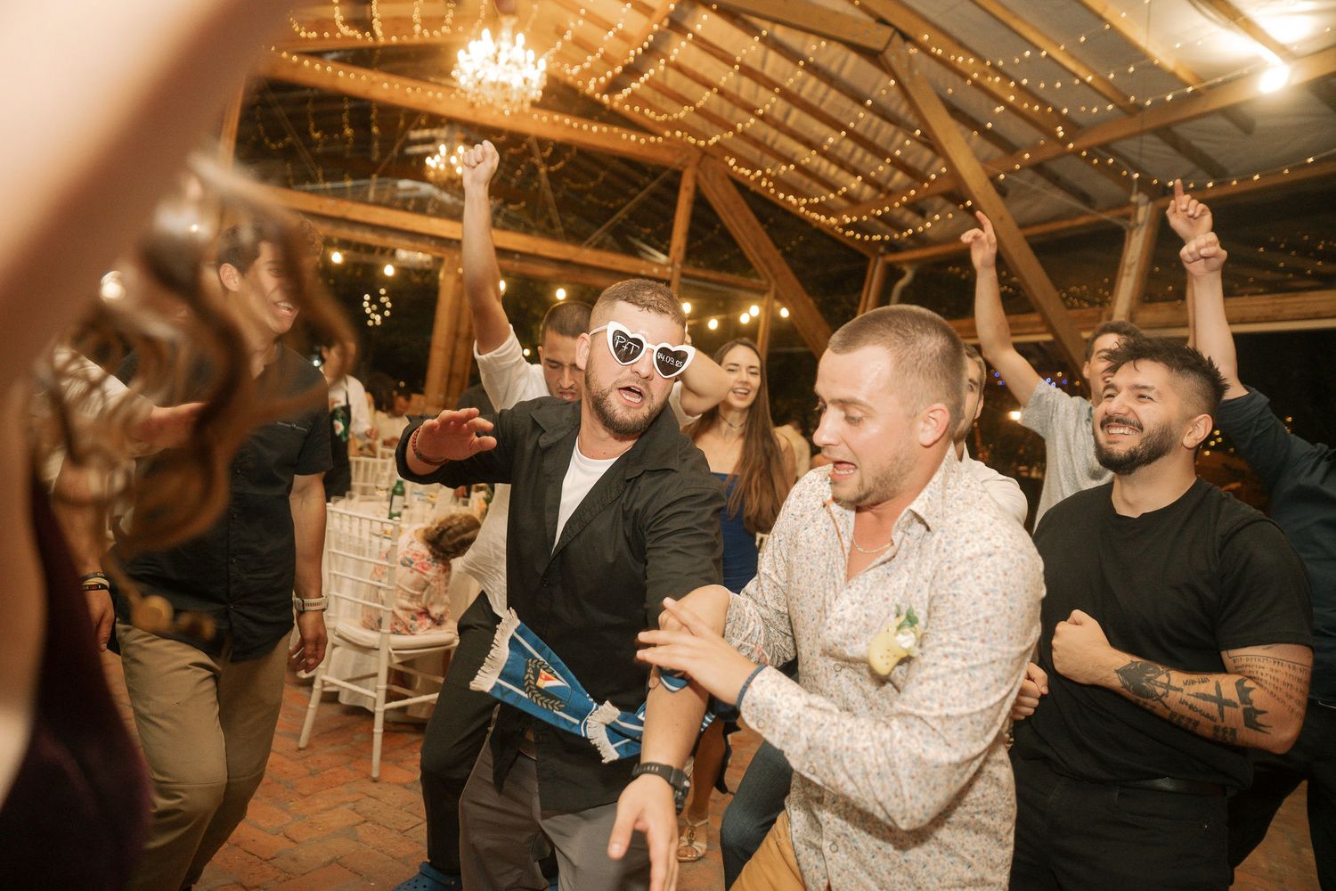 People dancing and celebrating at a lively indoor party with warm lighting and rustic wooden beams overhead.