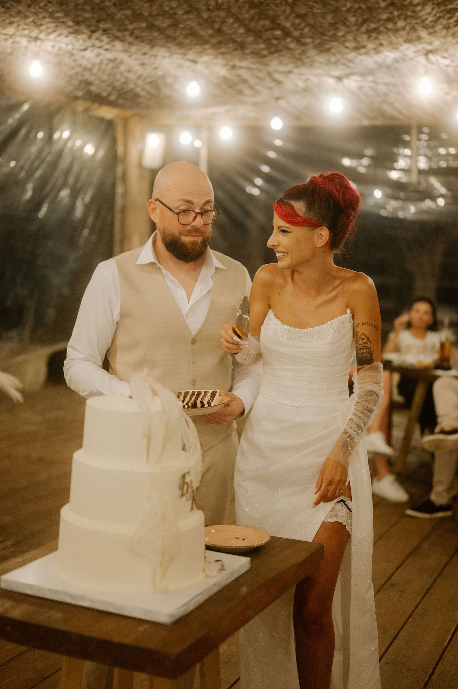 A couple in white attire cuts their wedding cake together under string lights at an evening reception.