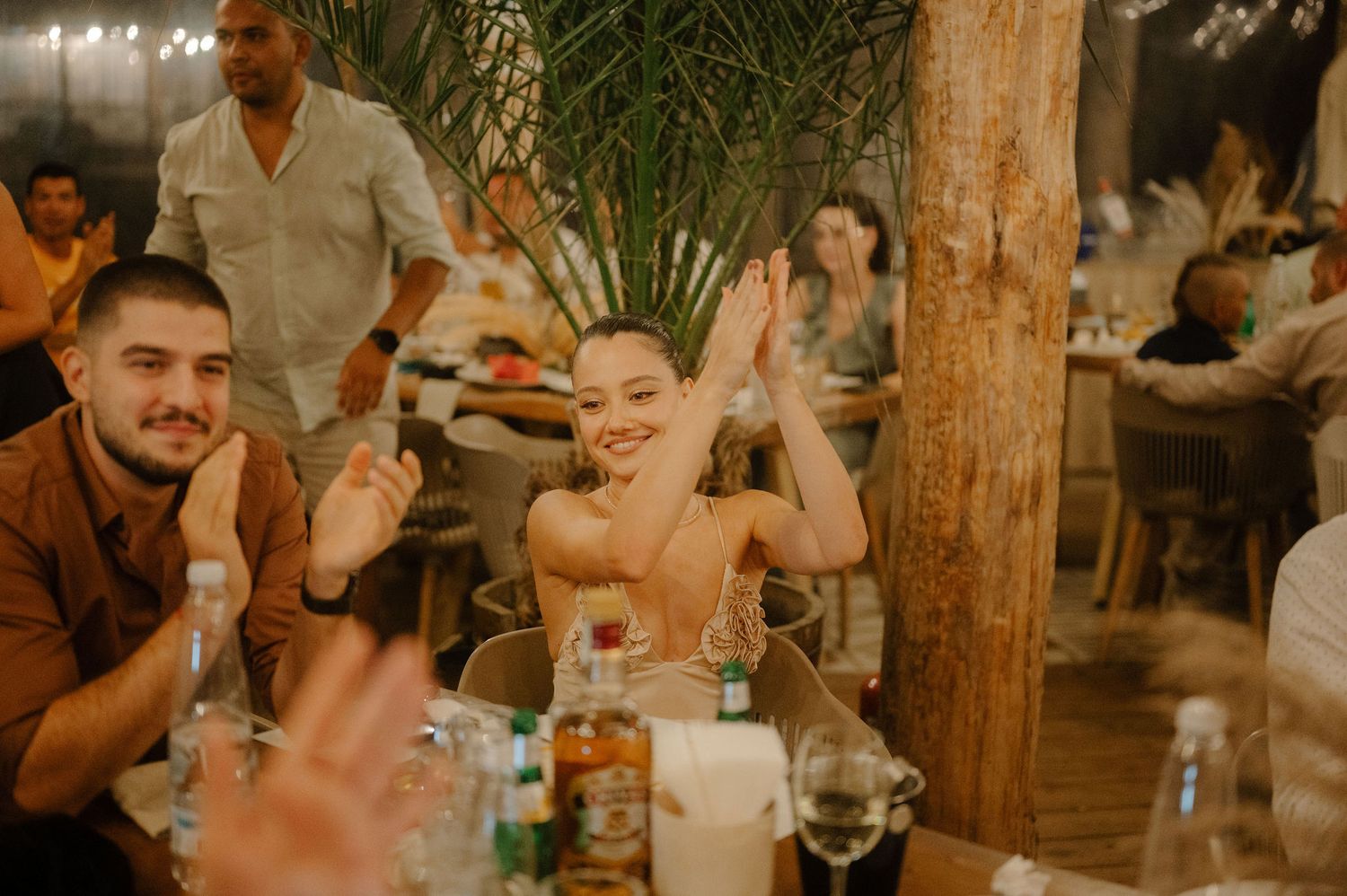 A group of people enjoying drinks and conversation at a rustic outdoor gathering with warm lighting and wooden decor.