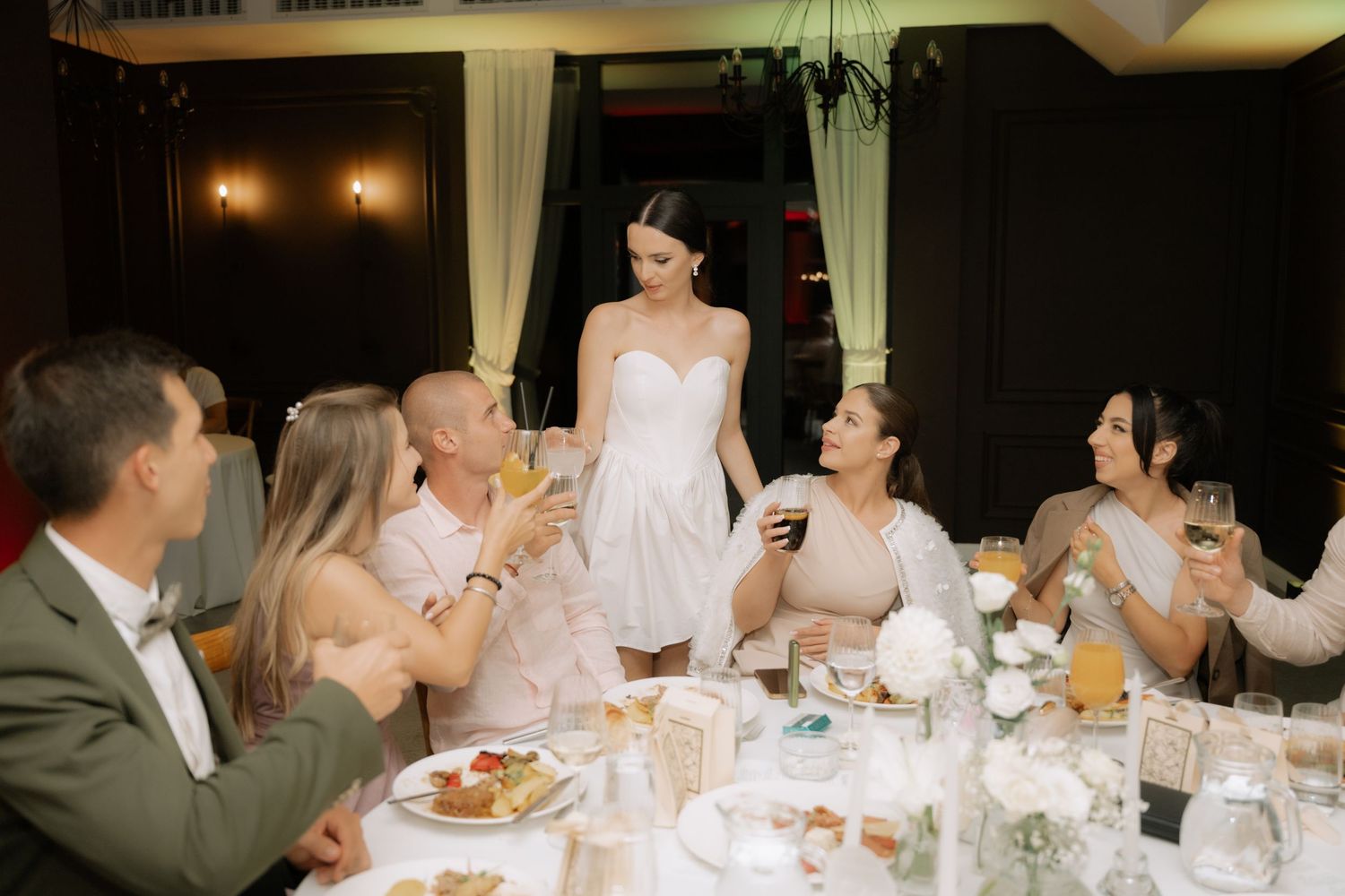 A group of wedding guests in formal attire laugh and enjoy a meal together at an elegant reception dinner table.