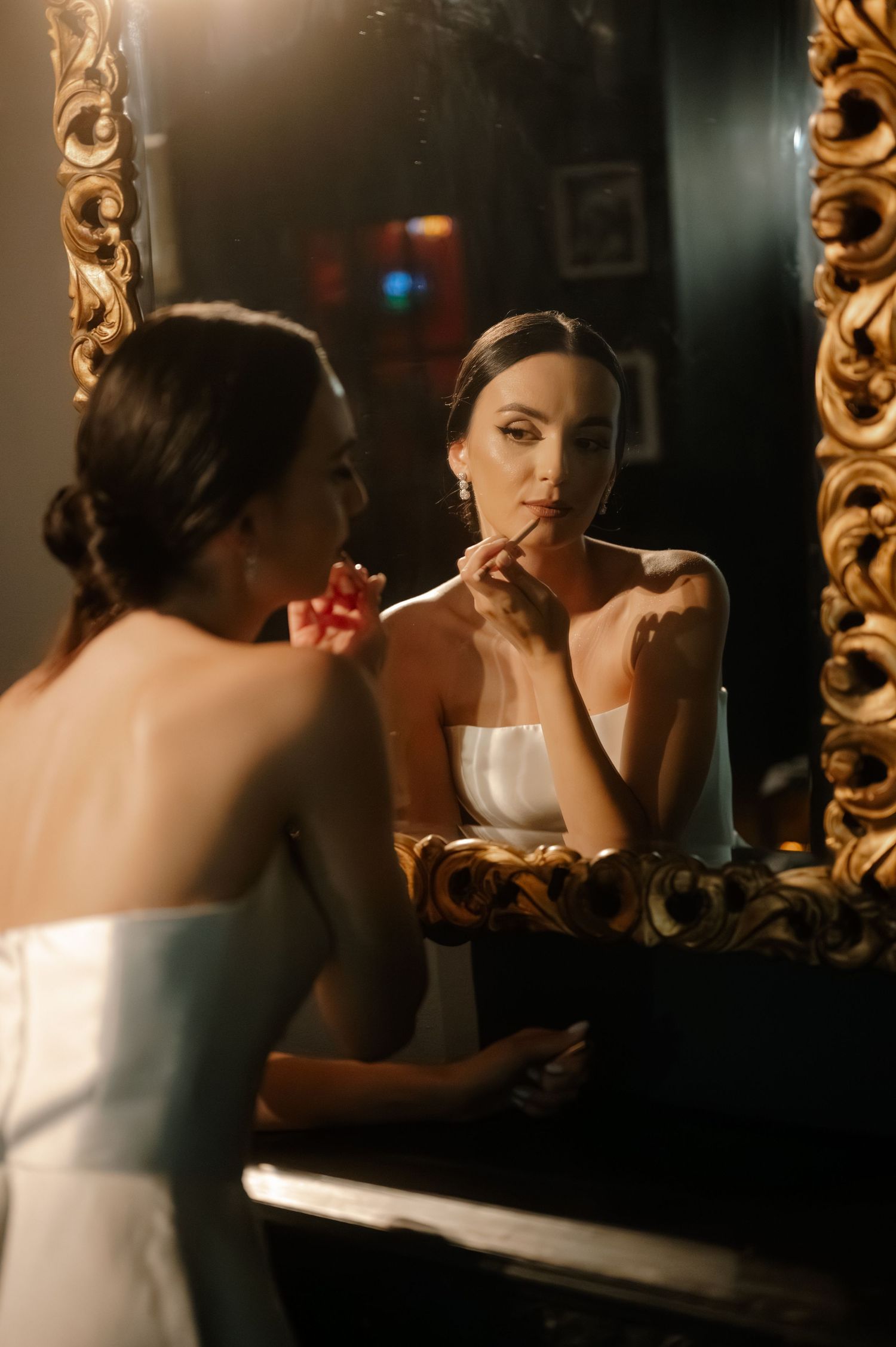 A woman in a white dress applies makeup while looking in a mirror surrounded by ornate gold frames.