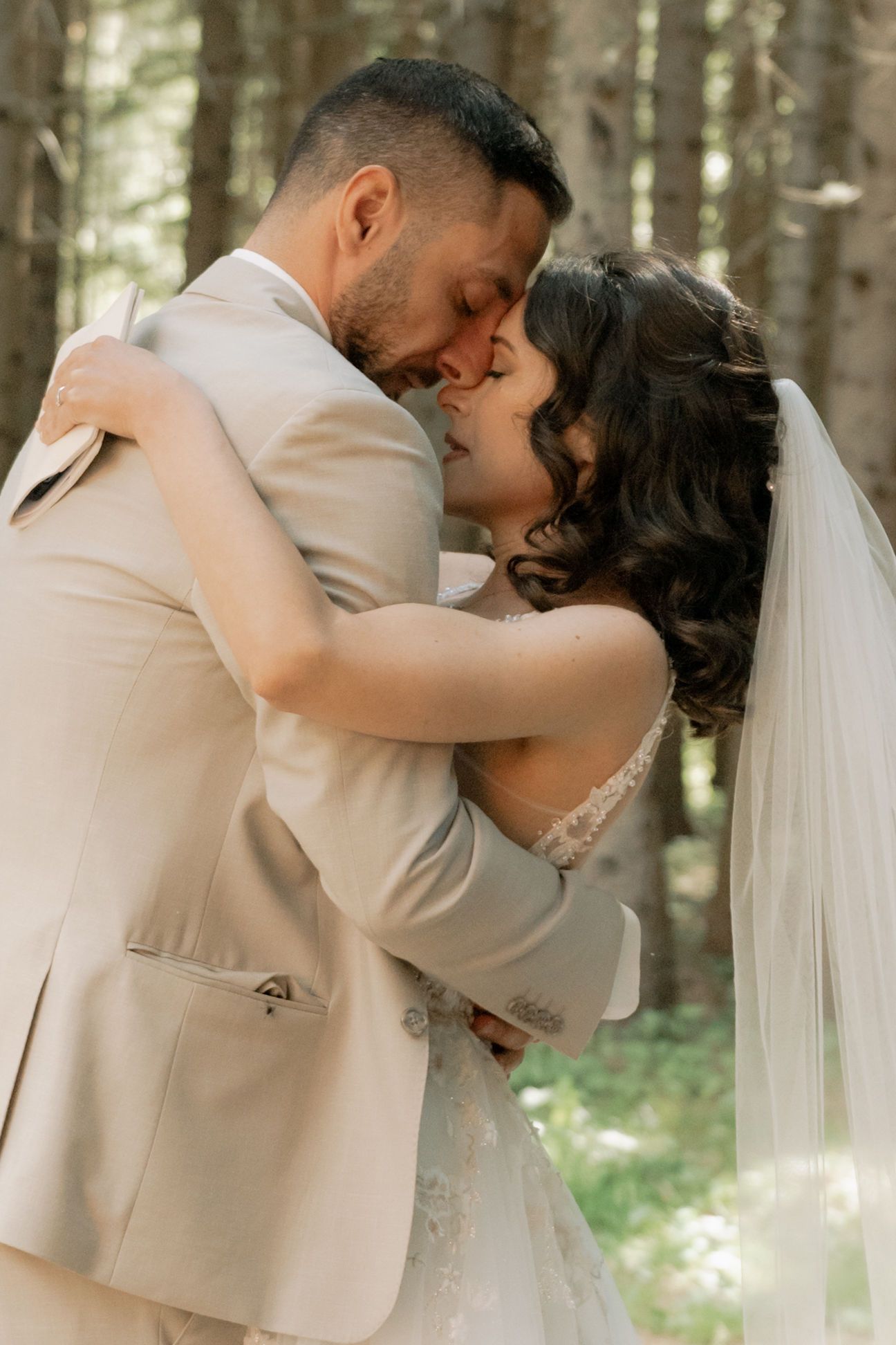 A bride and groom share a romantic embrace in a forest, wearing a white lace gown and beige suit on their wedding day.