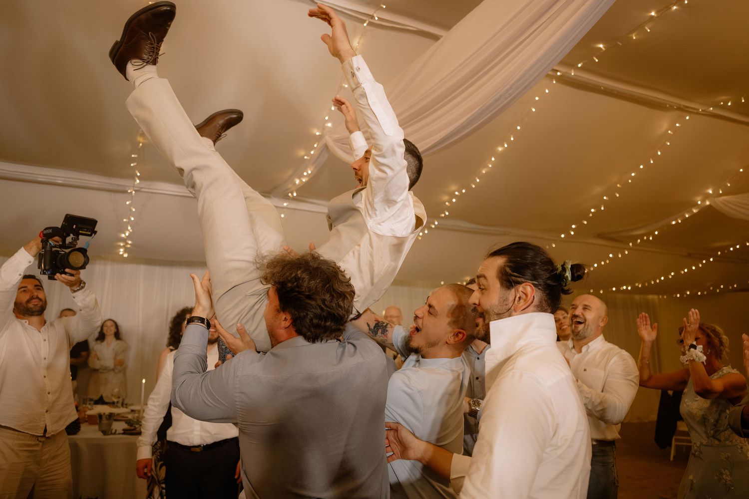 Wedding guests tossing a man into the air during an energetic reception party with fairy lights.