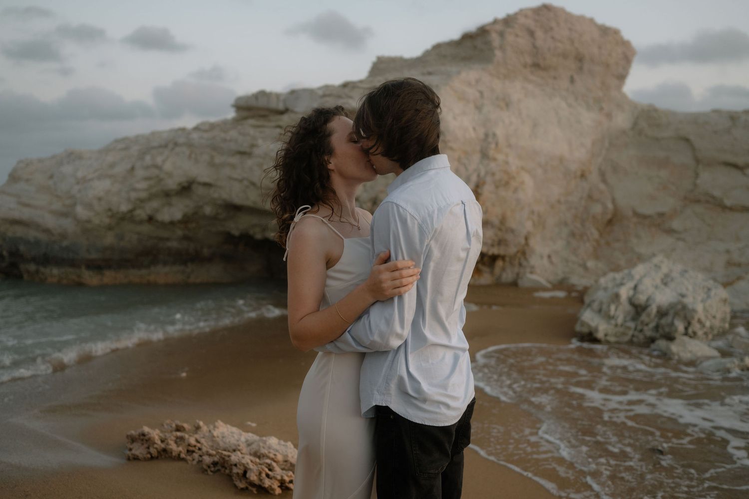 Couple sharing a romantic kiss on a sandy beach with dramatic rock formations and ocean waves in the background.