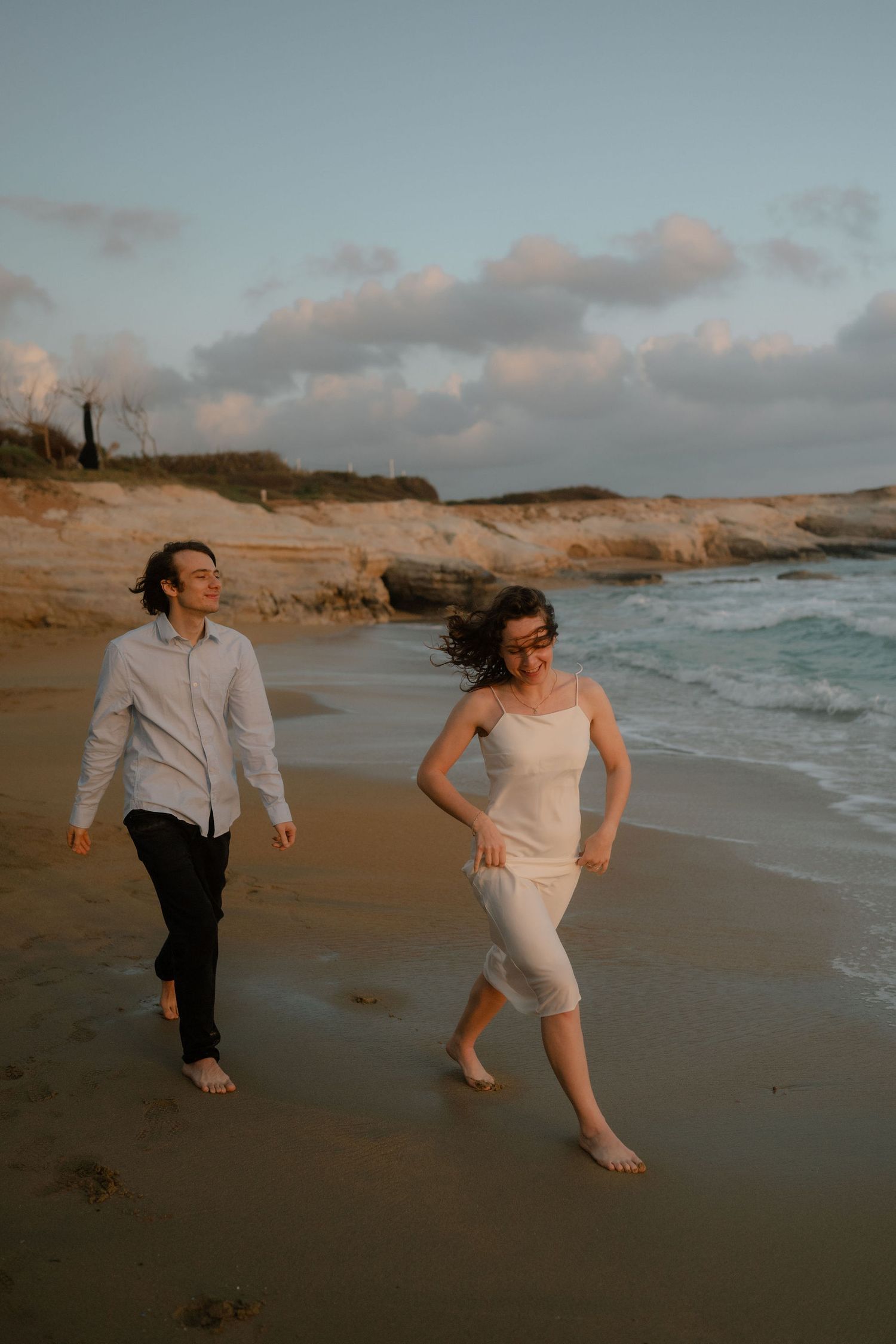 Couple walking barefoot on sandy beach at sunset, waves and rocky cliffs in background, wearing casual white outfits.