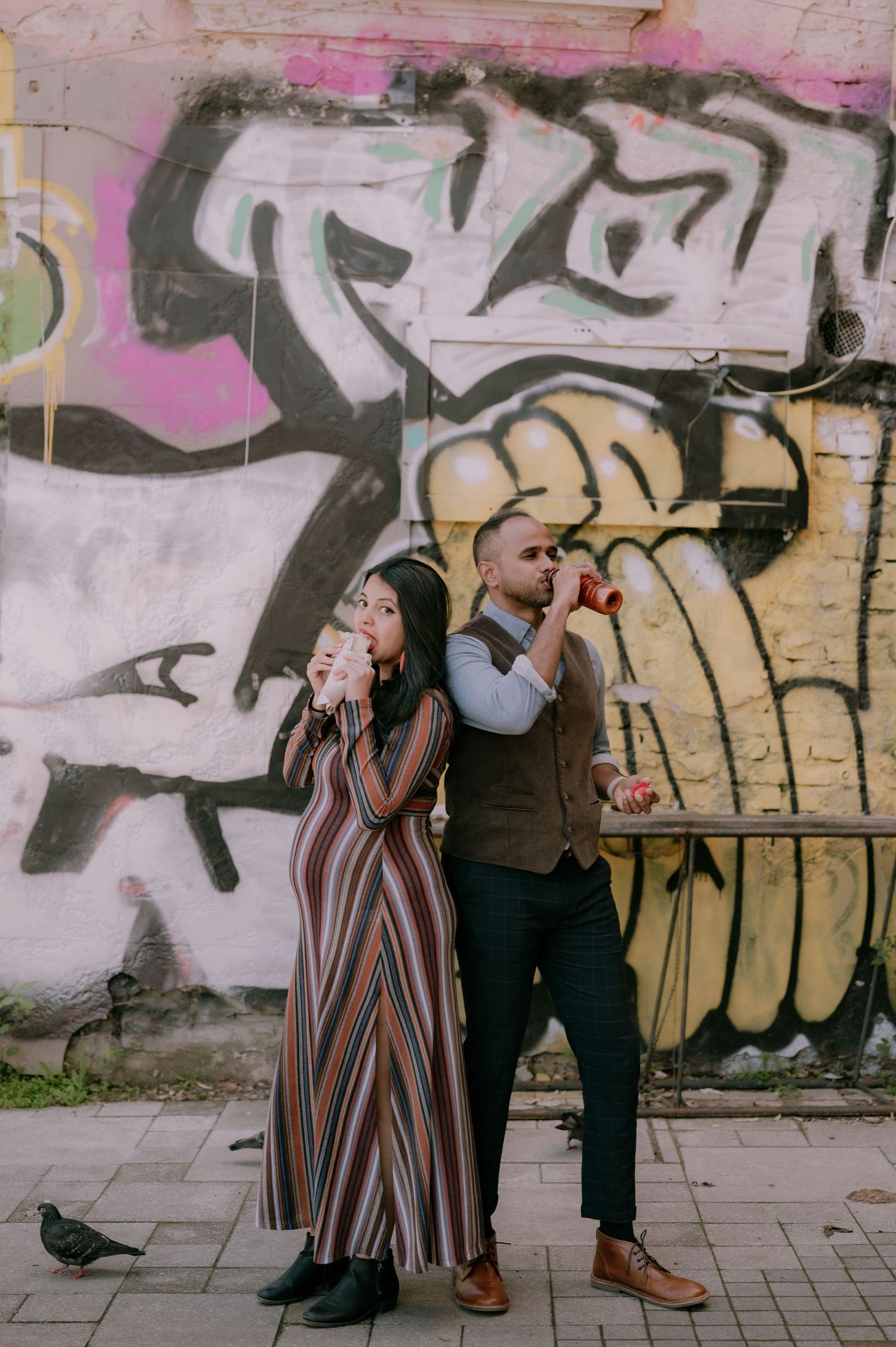 Couple posing back-to-back drinking beverages in front of colorful graffiti wall on urban street.