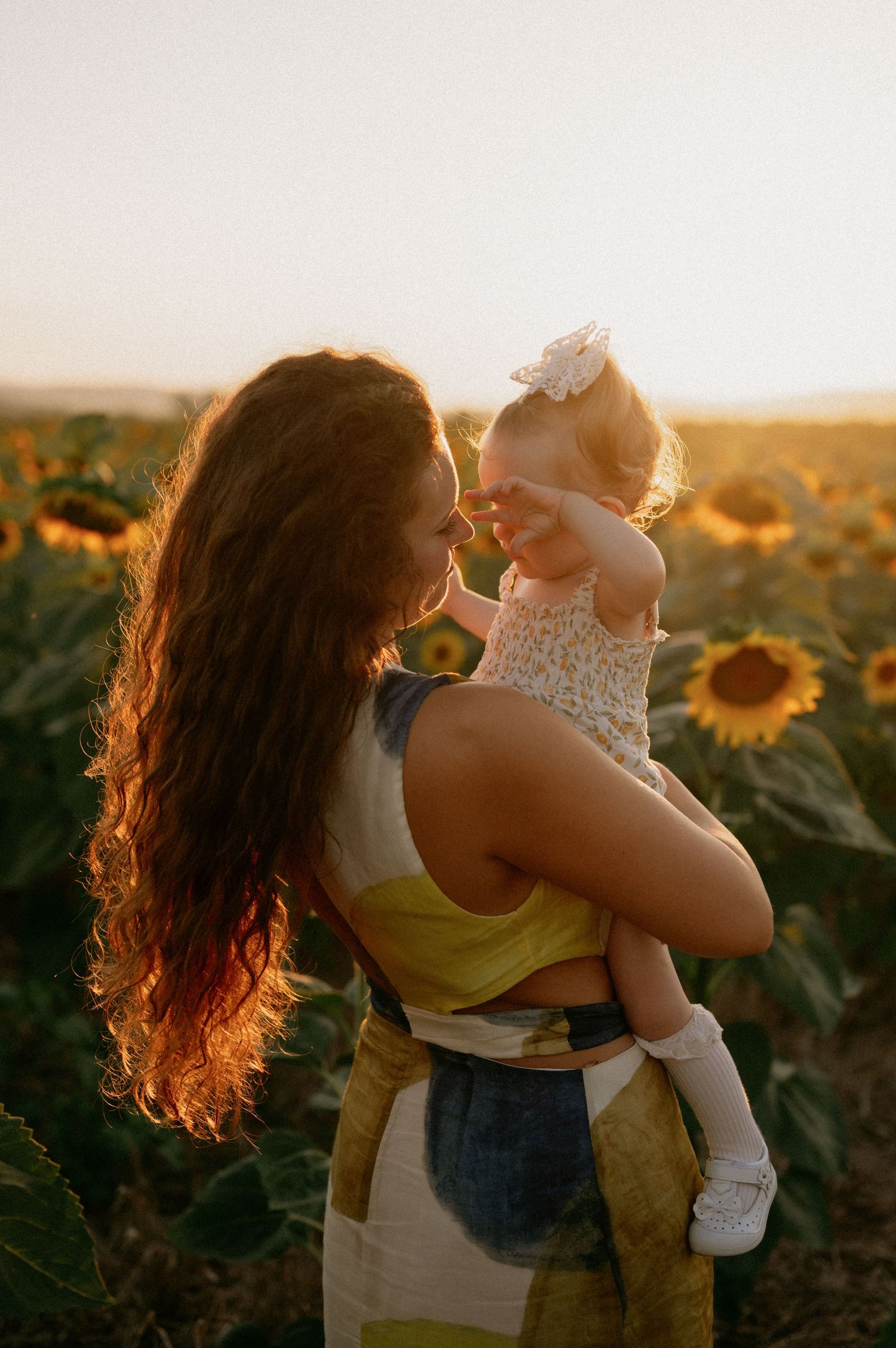 A mother holds her toddler in a sunflower field at golden hour, both sharing a tender moment in warm sunset light.