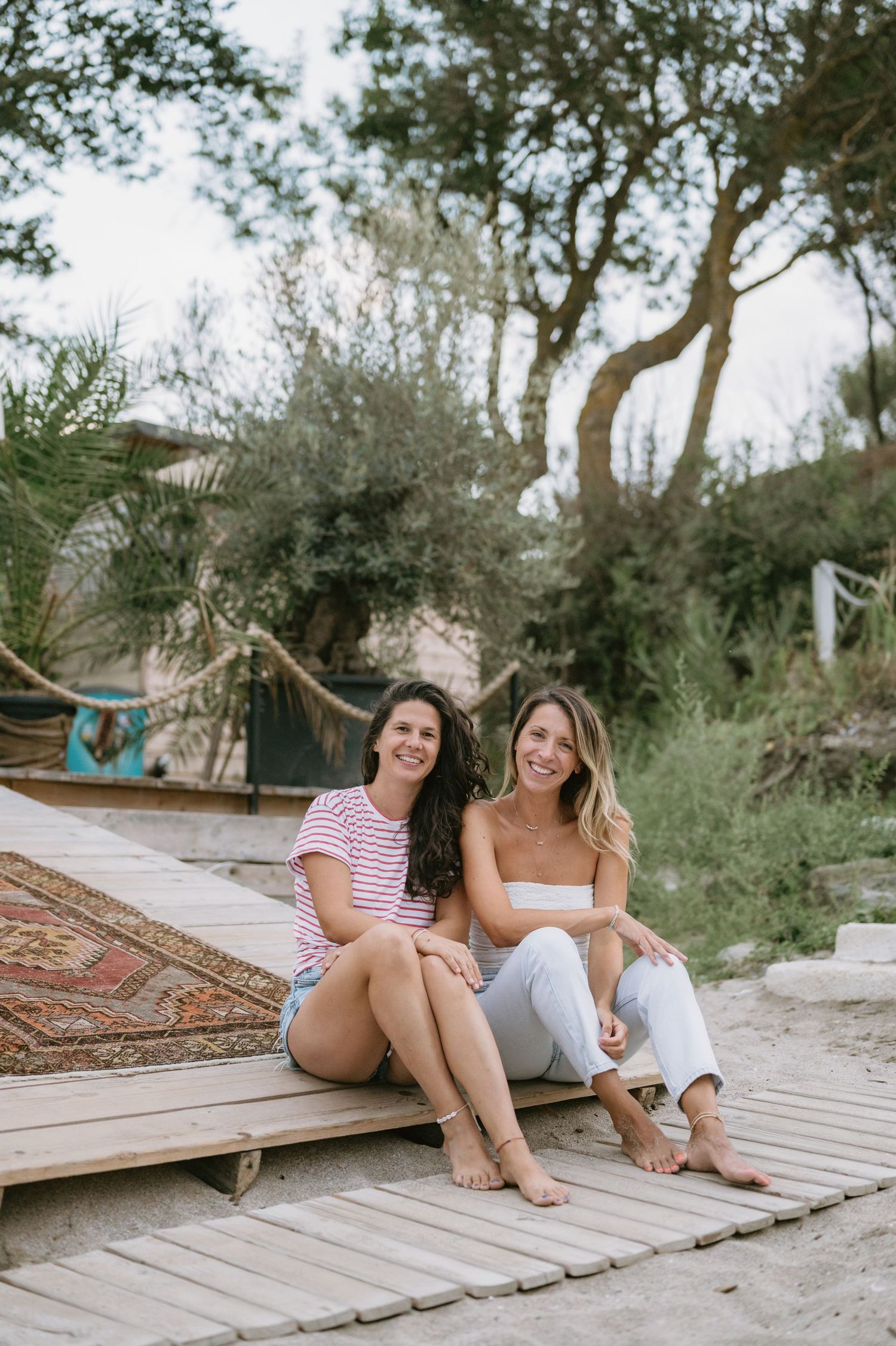 Two smiling women sitting together on a wooden boardwalk surrounded by trees and sand on a sunny day.