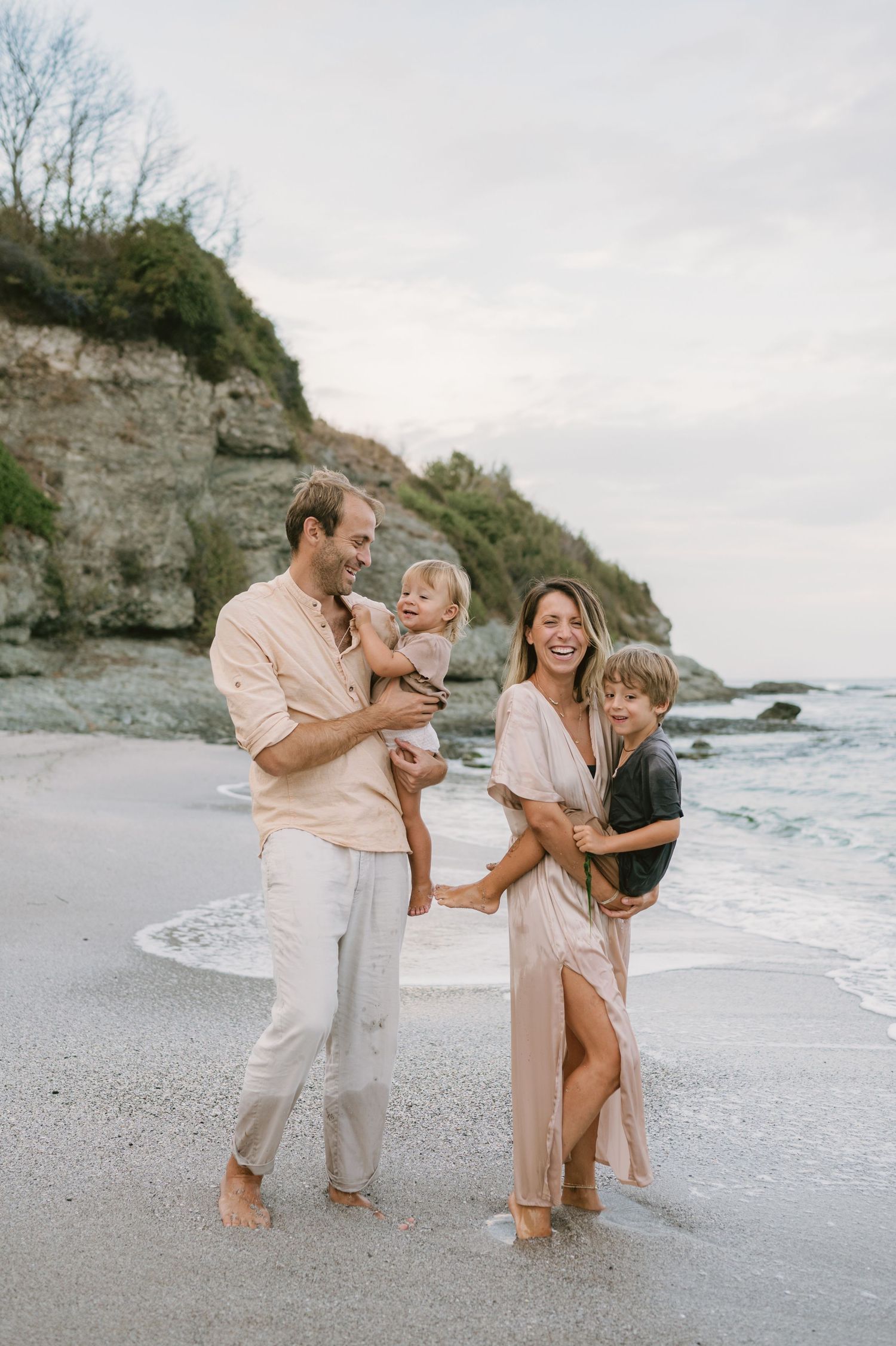 Happy family of four laughing together on a sandy beach, with rocky cliffs and ocean waves in the background.