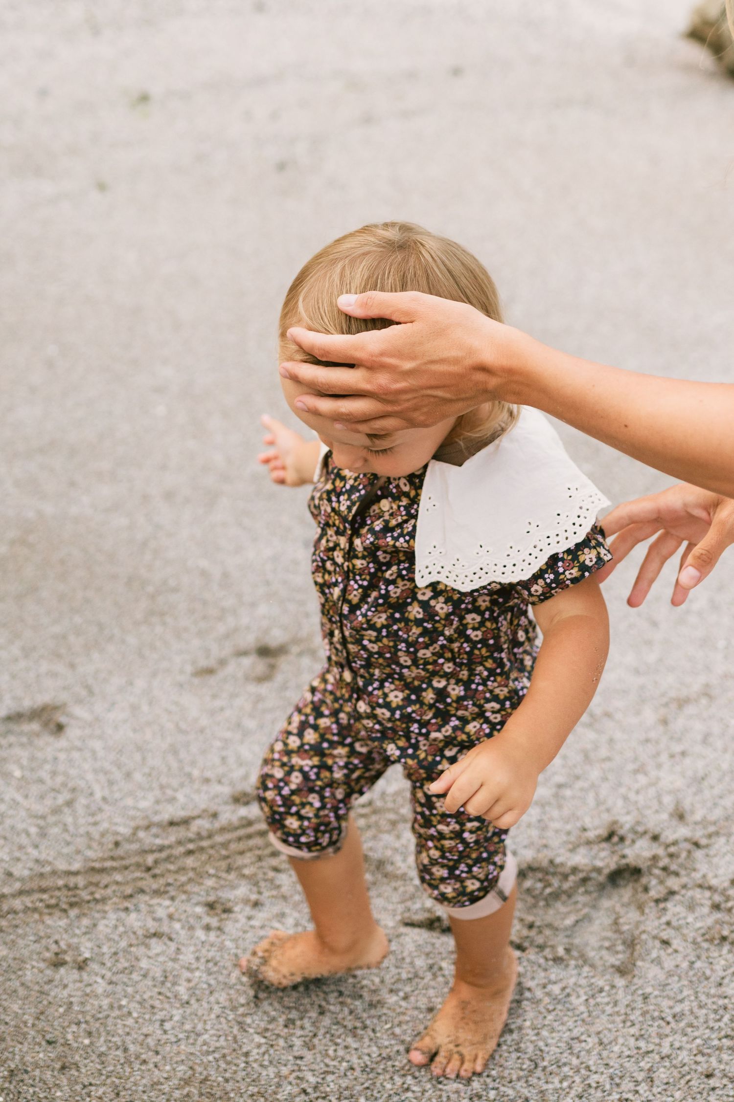 Toddler in floral romper with white collar walks barefoot on sandy beach, guided by an adult hand.