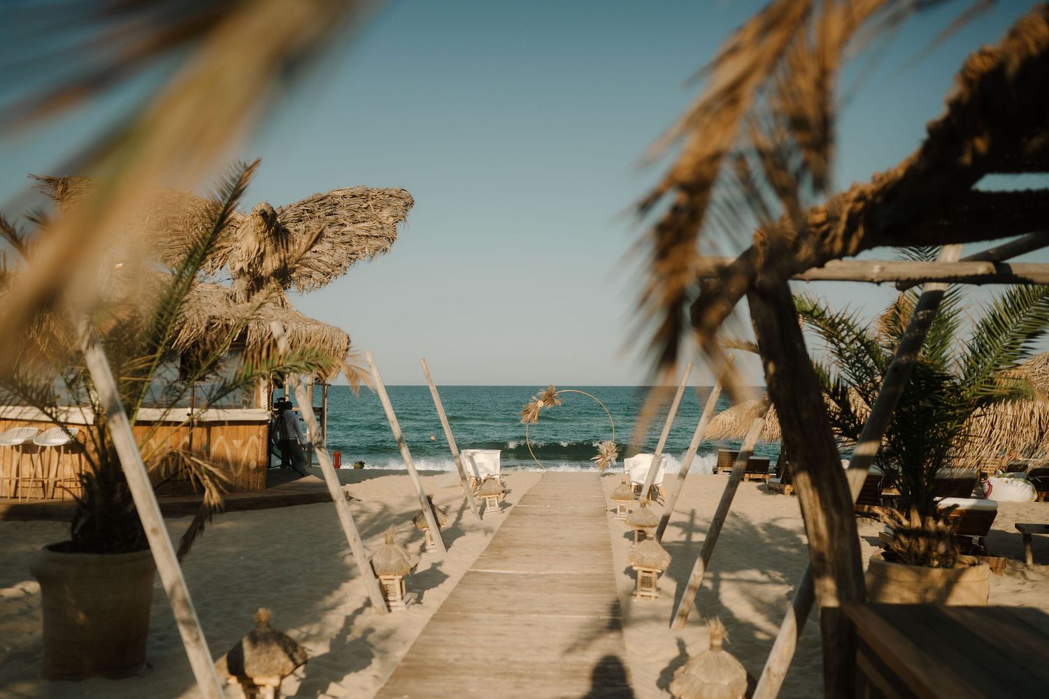 Wooden walkway leading to tropical beach with thatched umbrellas and white chairs framed by palm fronds.