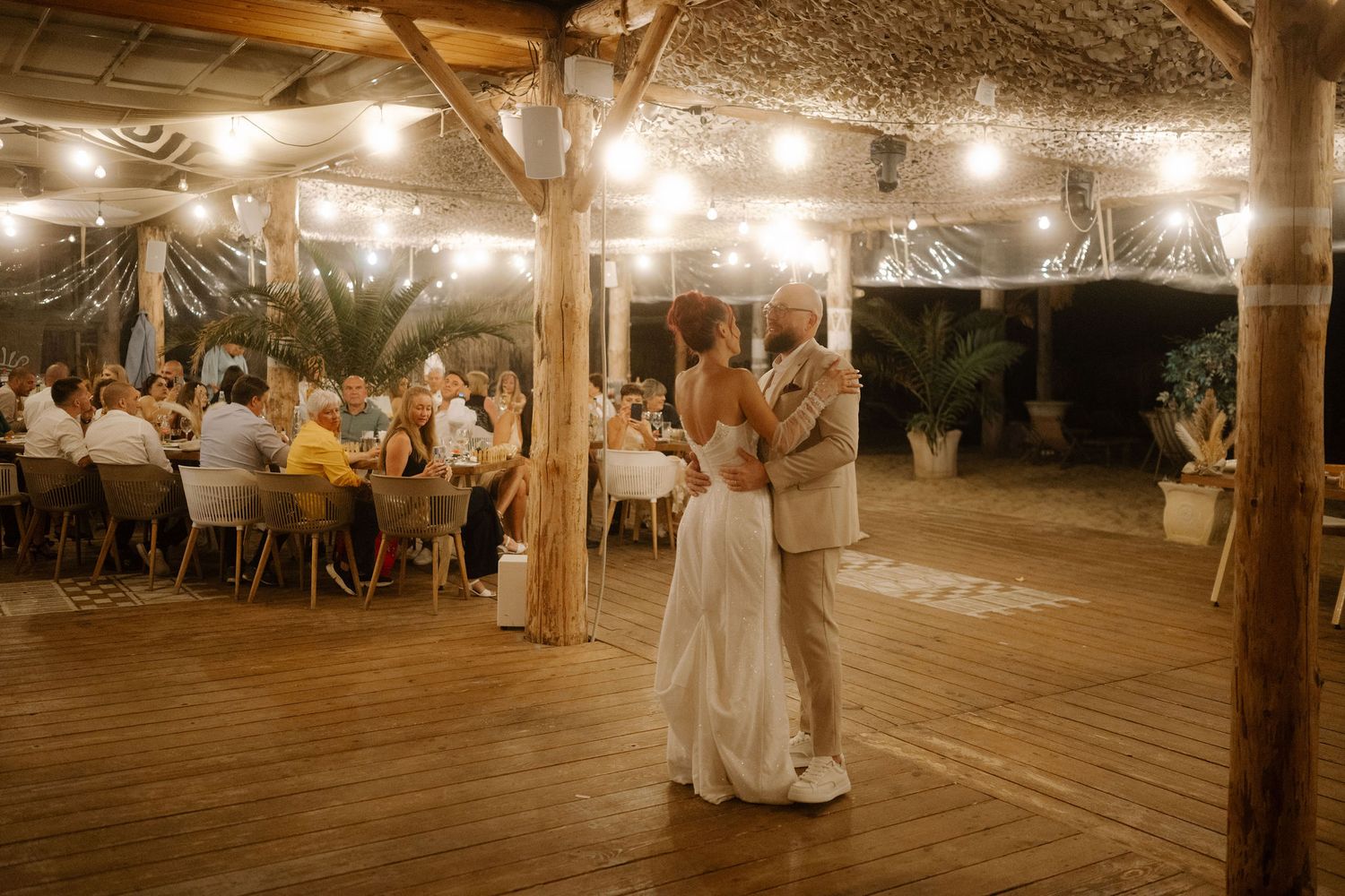 Wedding reception guests watching couple's first dance under string lights on outdoor wooden deck at night.