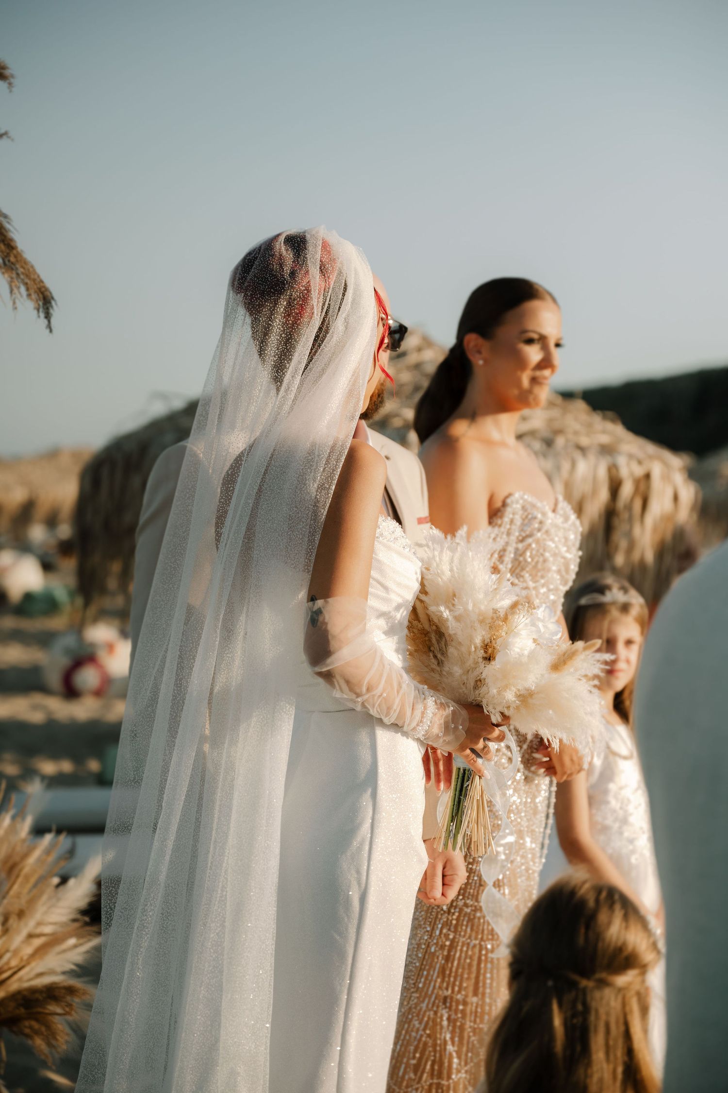 Elegant bride in lace wedding dress with flowing veil poses against scenic waterfront backdrop at sunset.