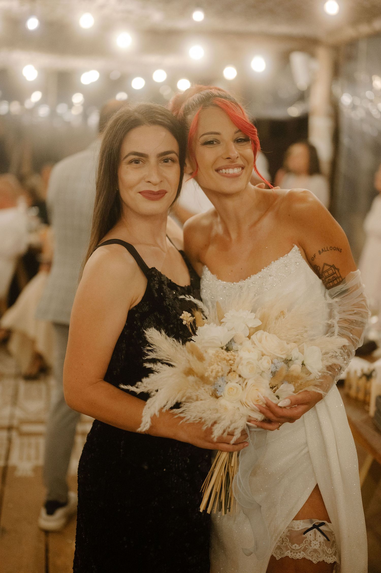 Wedding party members in black and white dresses pose together under warm string lights during an evening celebration.