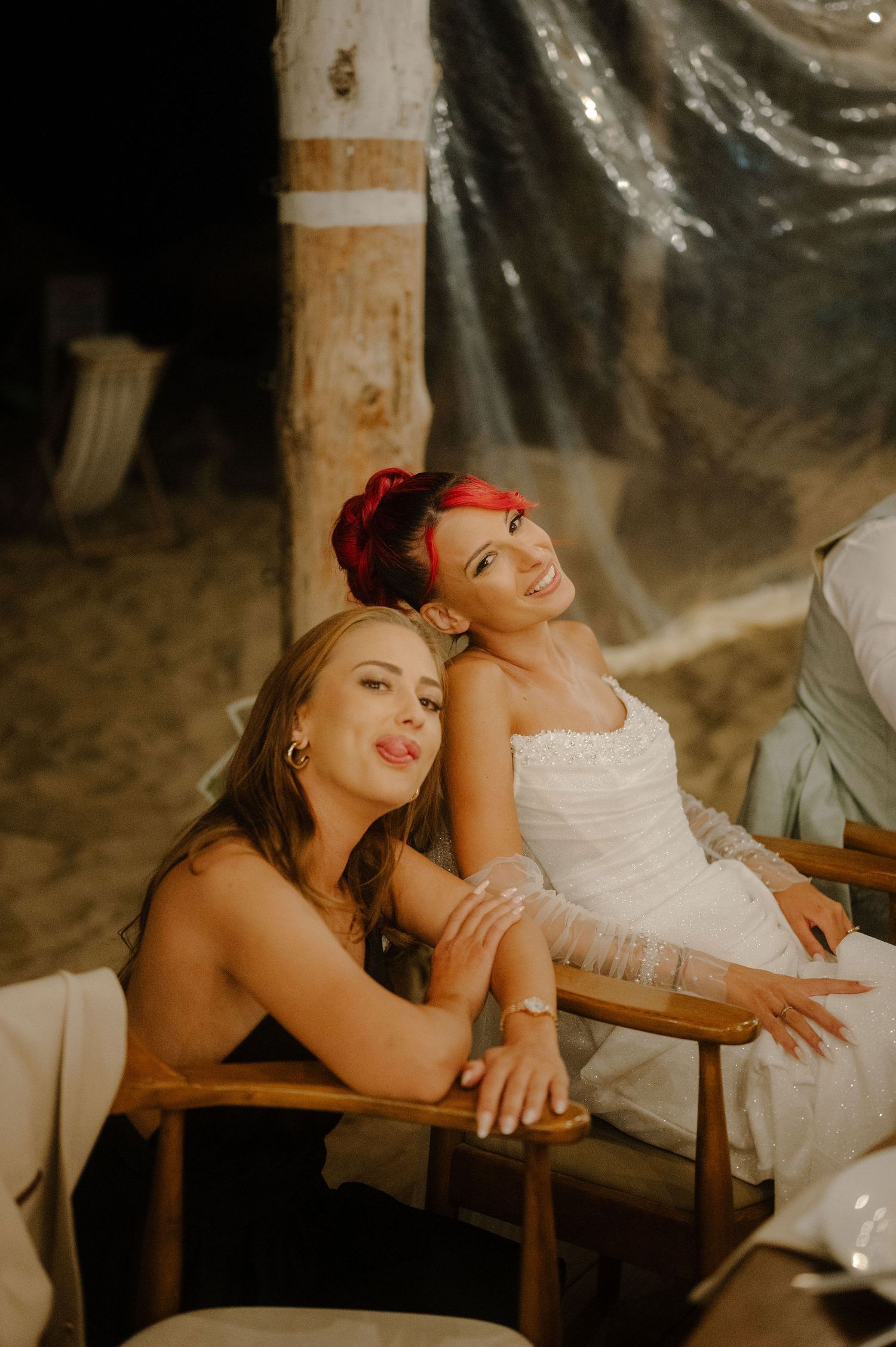 Two people in white dresses share a candid laughing moment while sitting together in a dimly lit space.