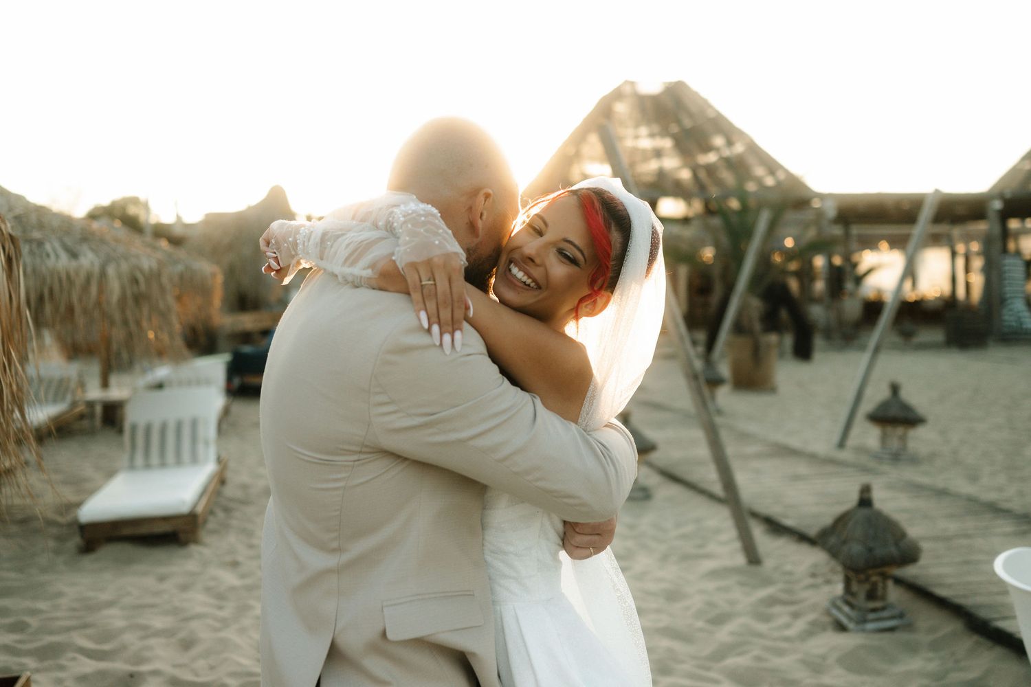 A romantic couple embraces during sunset on a beach with palm trees and lounge chairs in the background.
