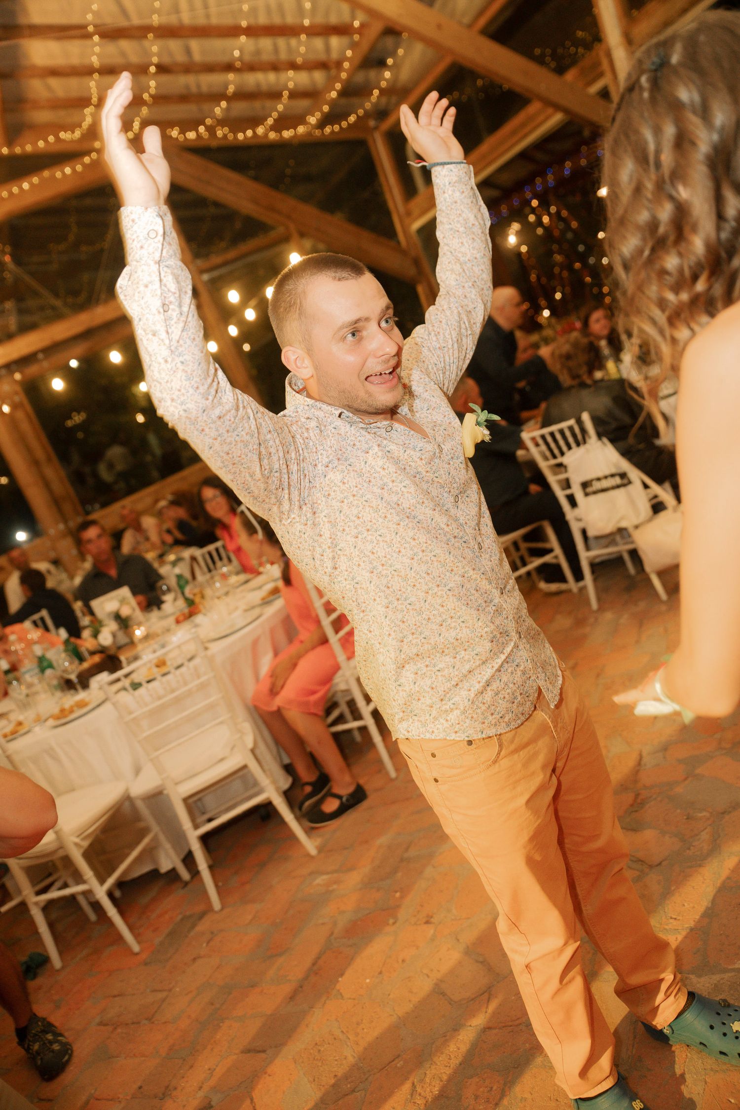 Energetic wedding guest in beige shirt and khaki pants dances enthusiastically on wooden dance floor under string lights.