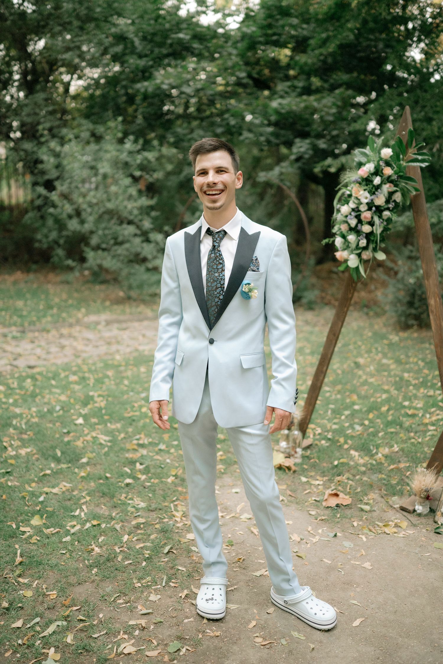 A person in a light gray suit and polka dot tie walks on a leaf-covered path in an outdoor garden setting.