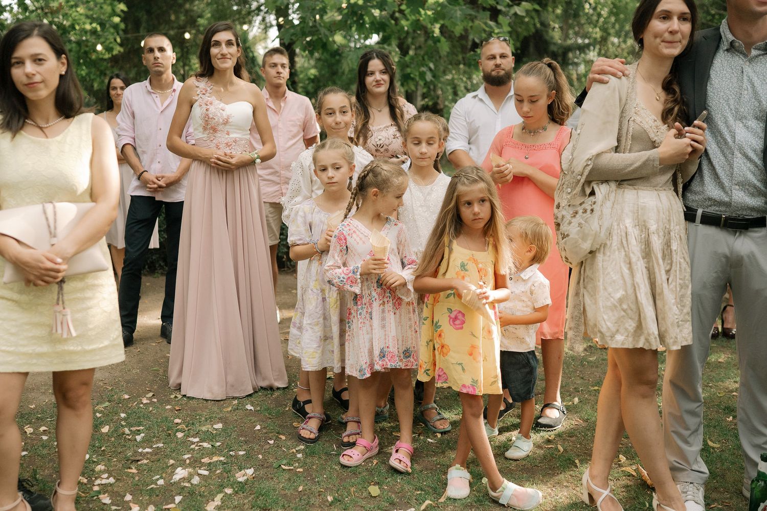 Wedding party members in pink and peach dresses gather outdoors with young children in matching attire.