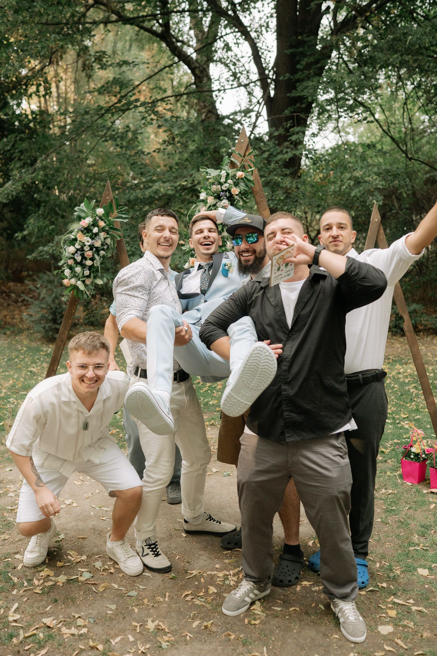 A group in light colored clothing poses playfully in a wooded outdoor setting during a wedding celebration.