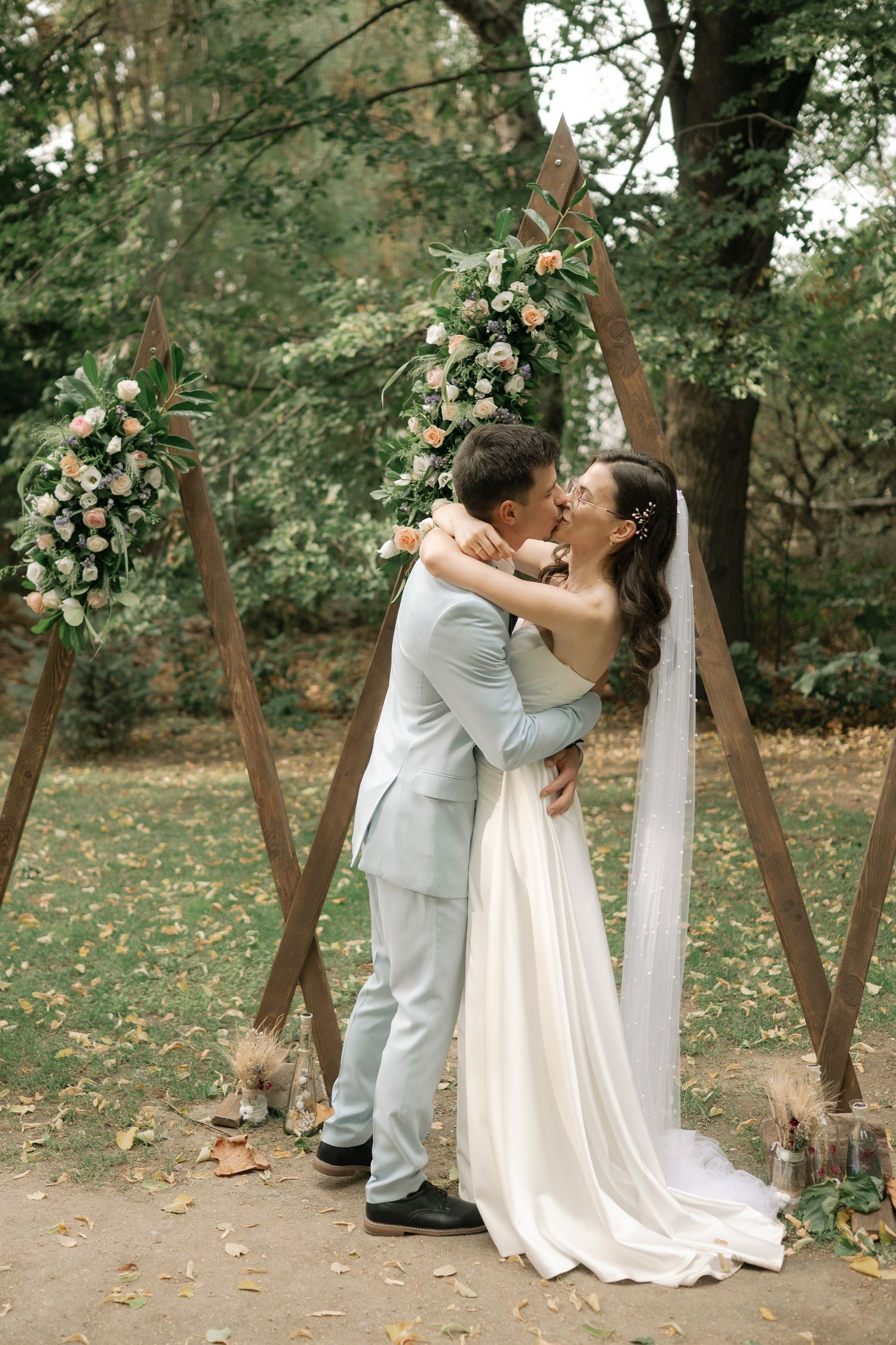 A romantic wedding couple shares intimate kisses in a wooded outdoor setting with autumn leaves on the ground.