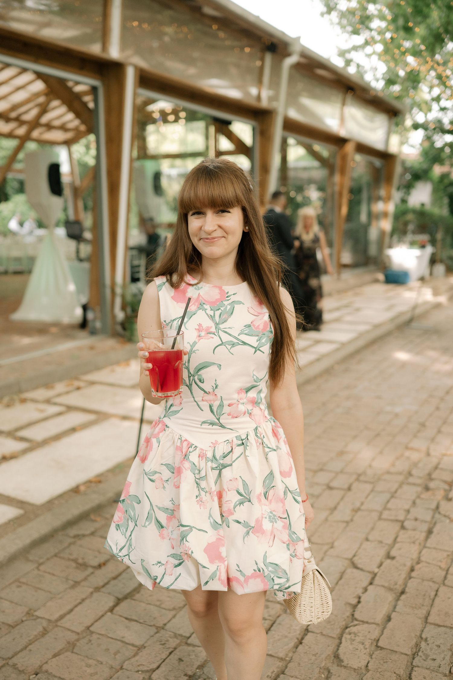 A person in a floral print dress stands on a brick walkway holding a red beverage on a sunny day.