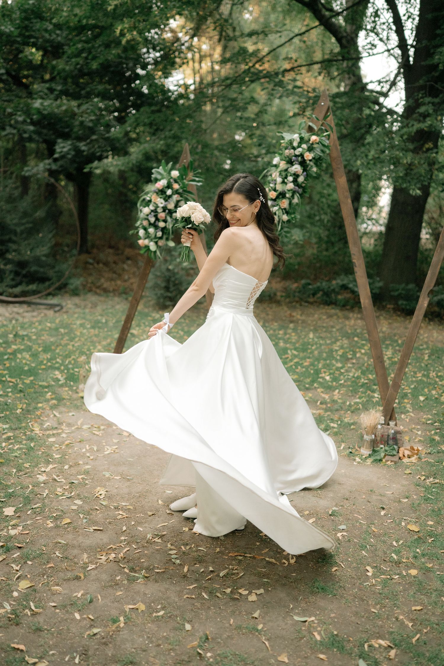A bride in a flowing white wedding dress twirls gracefully in a wooded garden setting with floral decorations.
