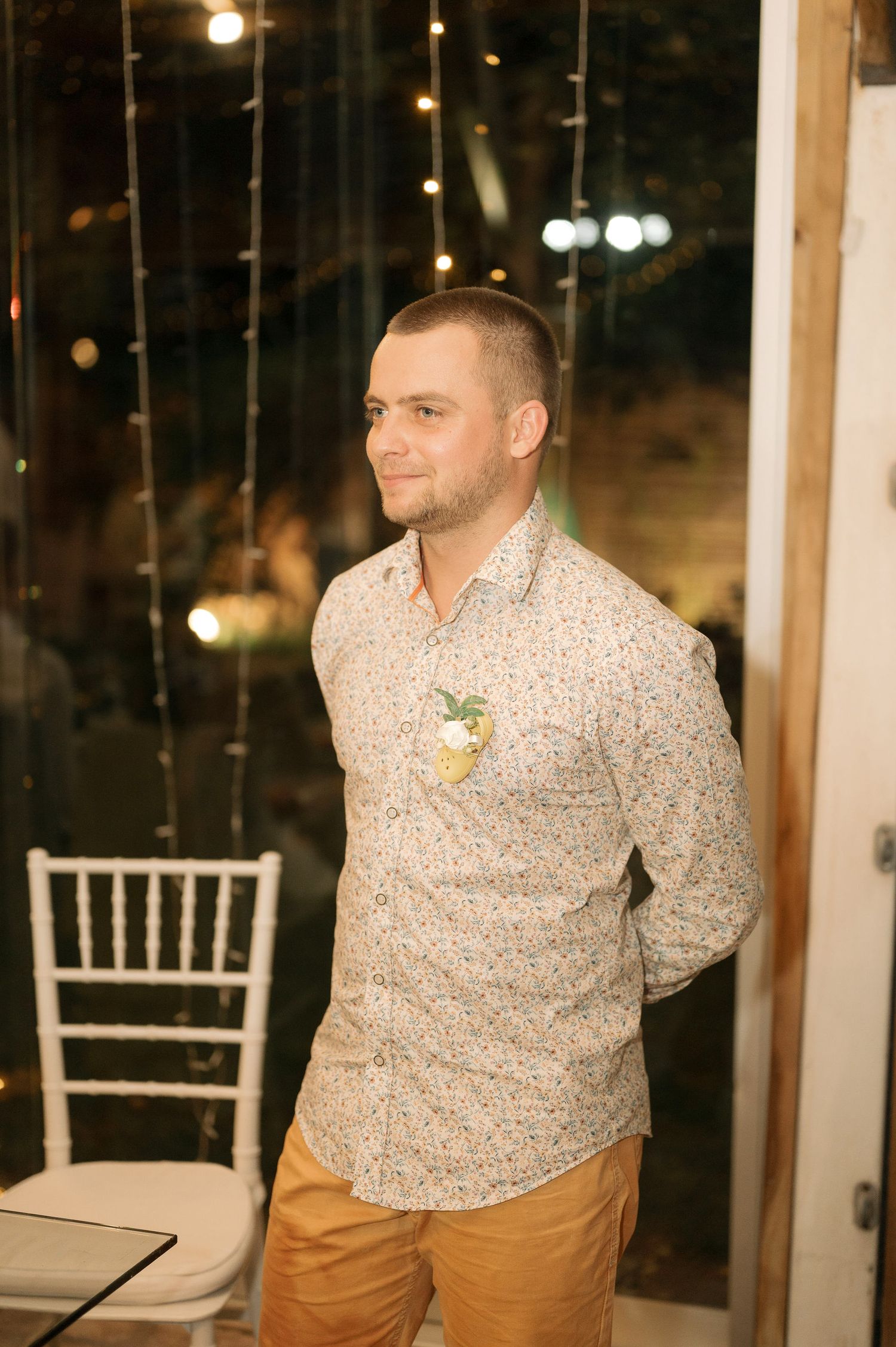 A person in a patterned button-down shirt and khaki pants stands at an indoor event with string lights and white chairs.
