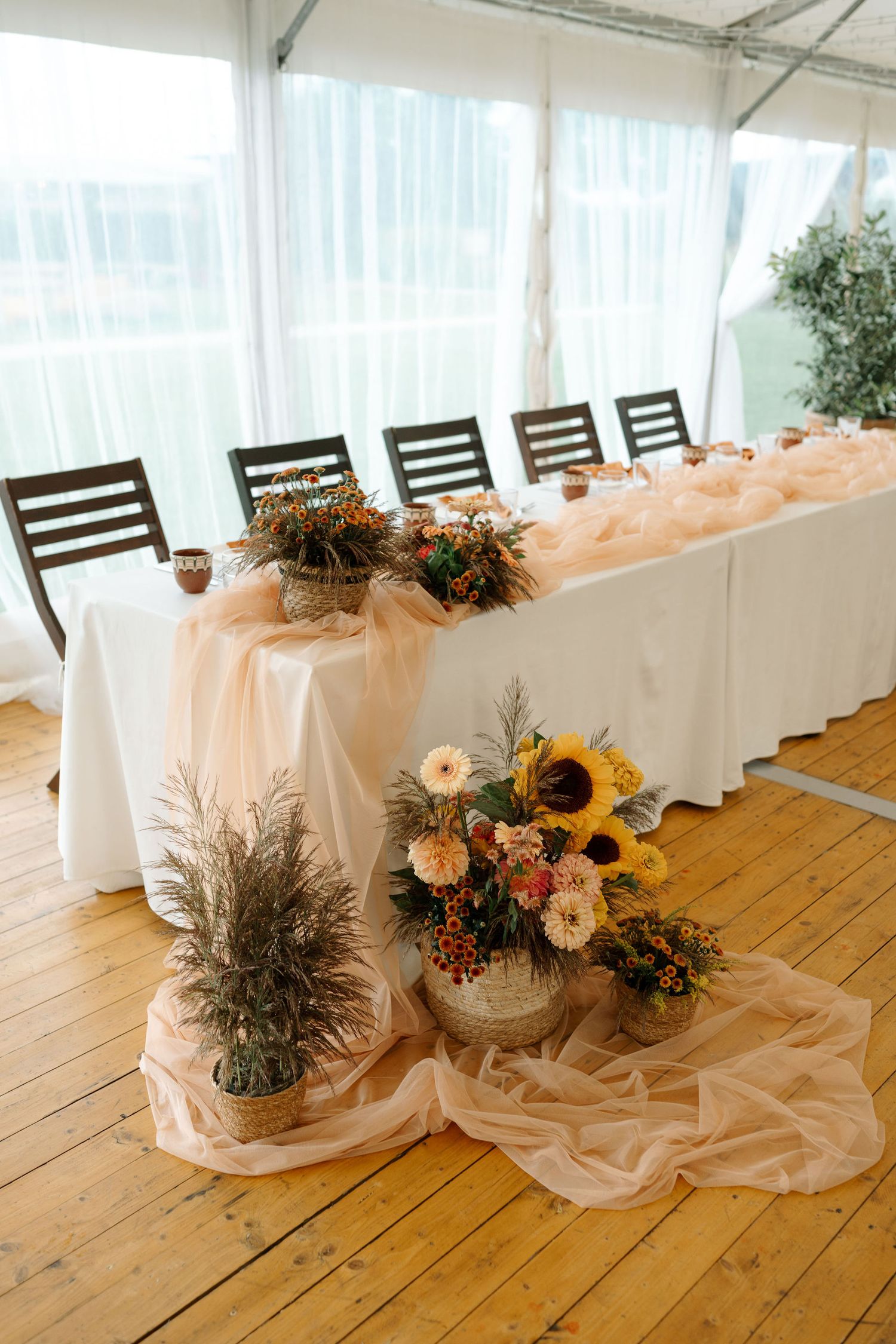 Rustic wedding table decor with sunflowers and floral arrangements on white tablecloths in a tent venue with wooden floors.