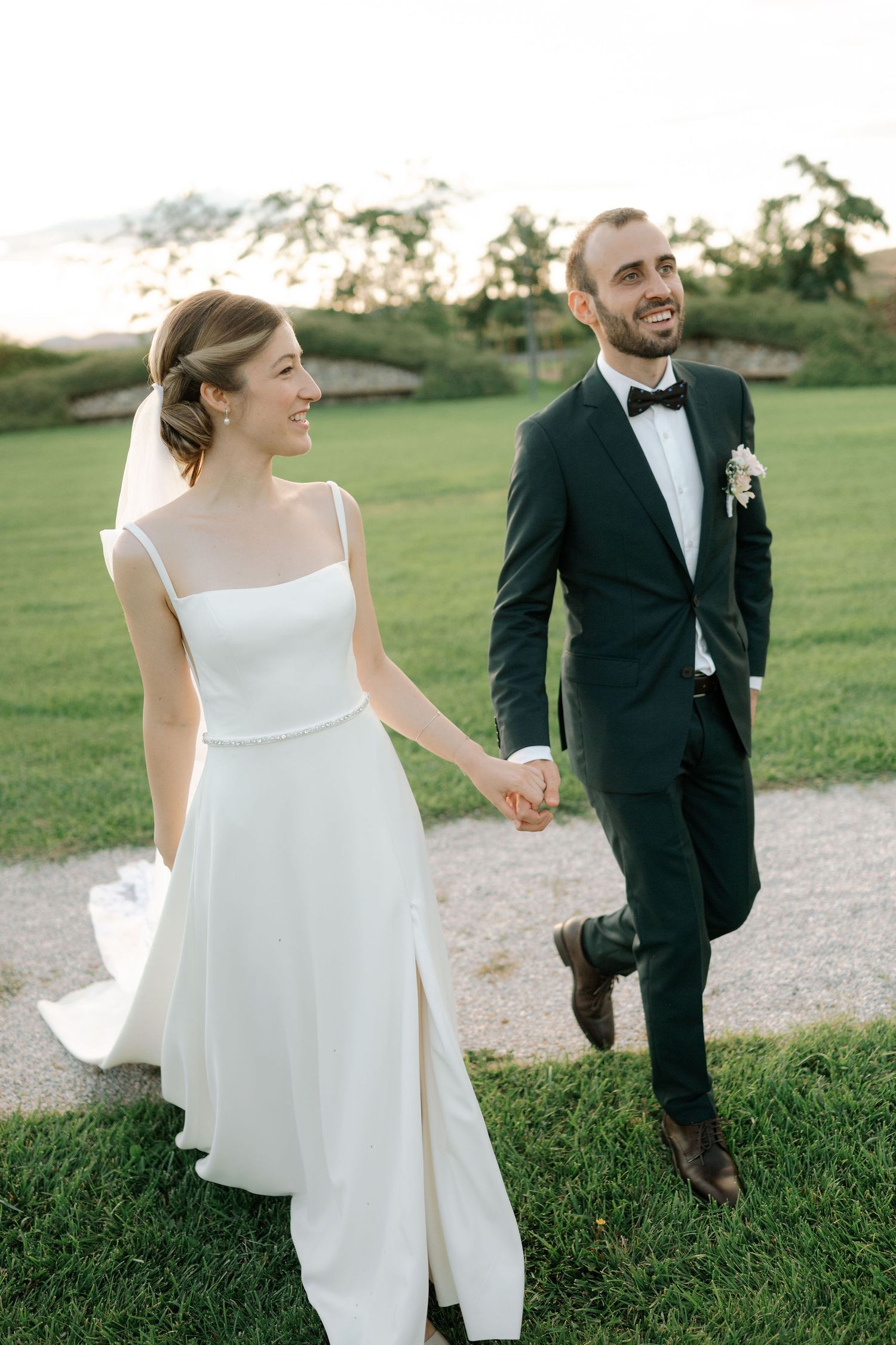 A bride and groom walk together on a grassy path during their outdoor wedding photoshoot at sunset.
