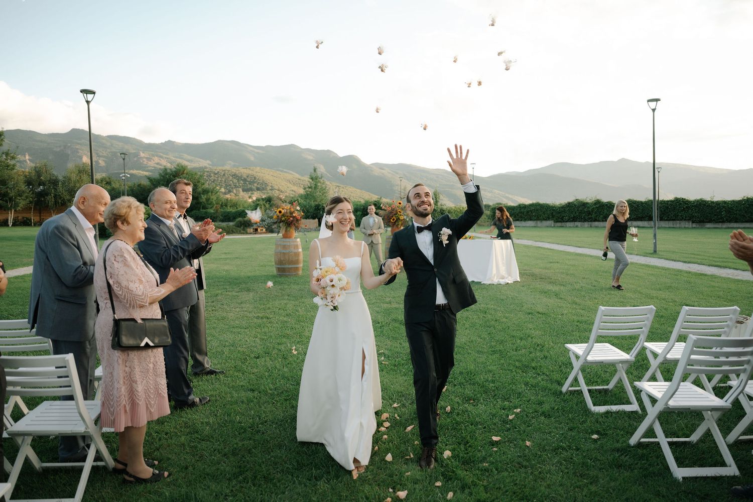 Wedding celebration on a lush green lawn with confetti flying as guests cheer at an outdoor ceremony.