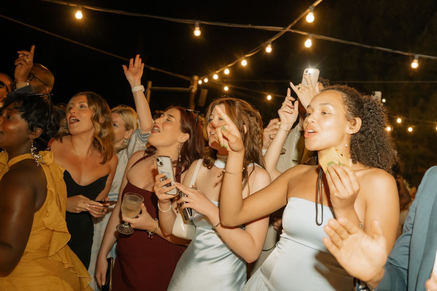 Women dancing and celebrating at an outdoor wedding reception under string lights at night.