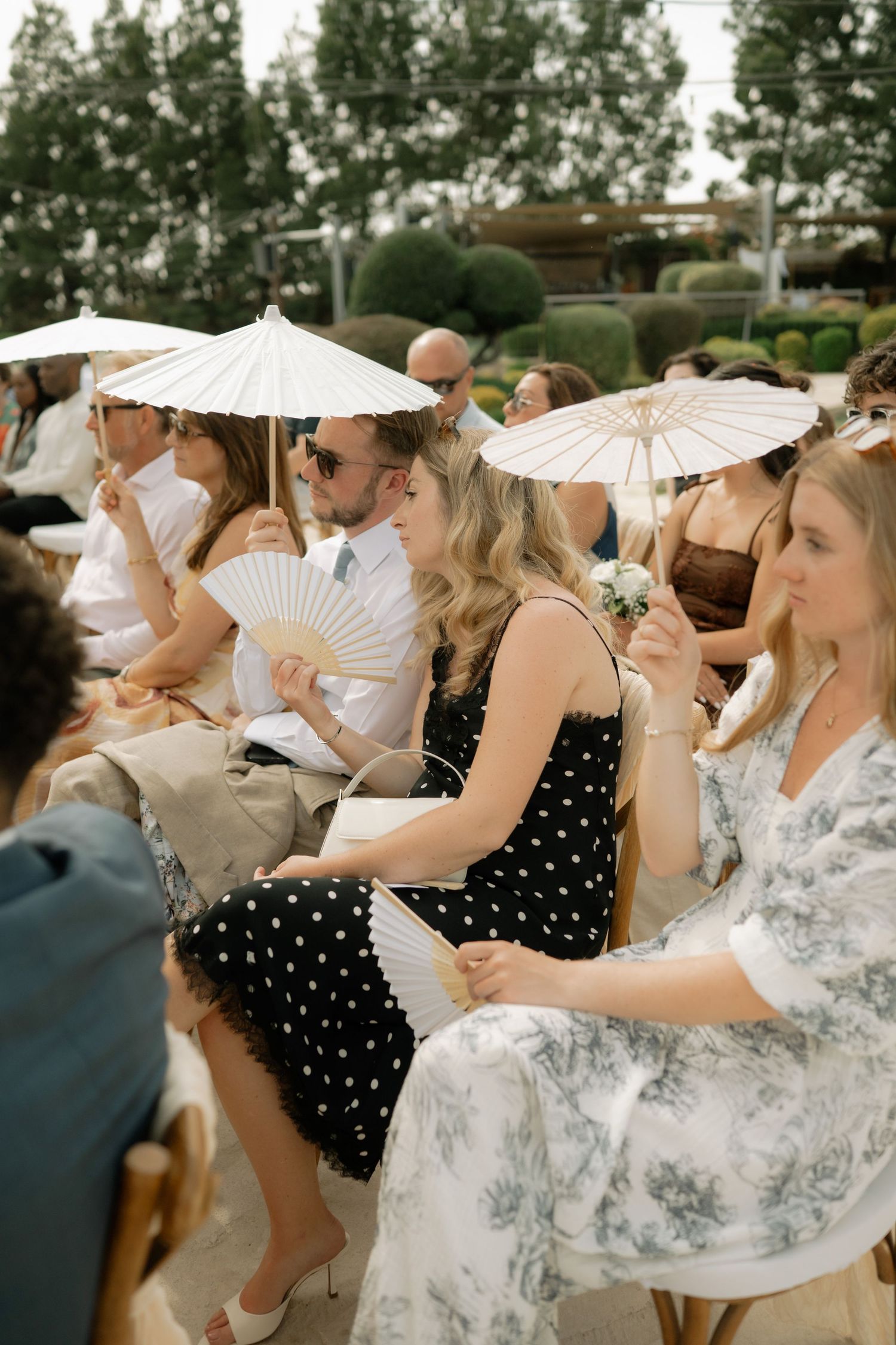 Wedding guests seated outdoors holding white parasols and fans, dressed in elegant summer attire at a garden ceremony.