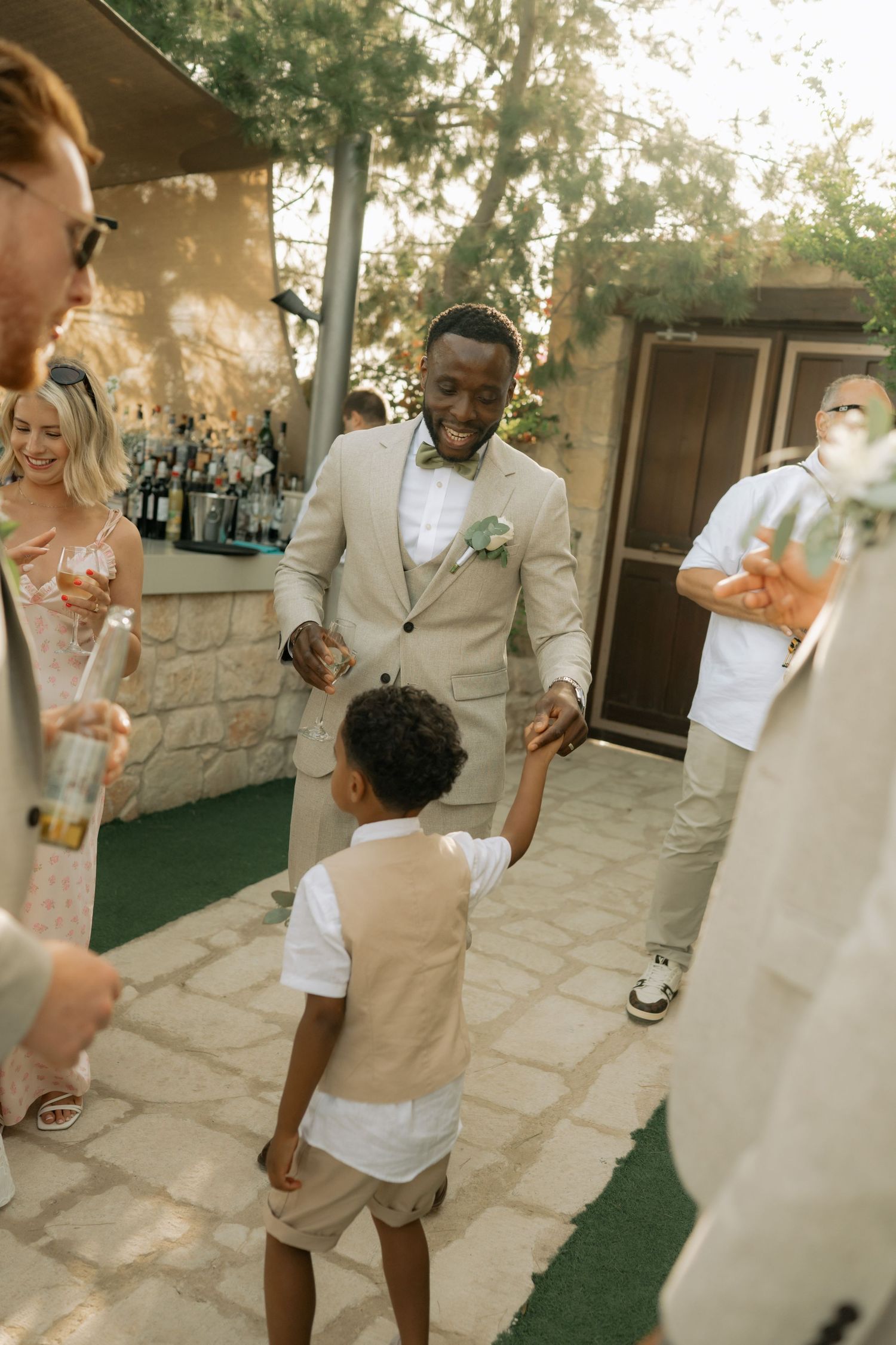A groom in a beige suit dances joyfully with a young boy at an outdoor wedding reception.