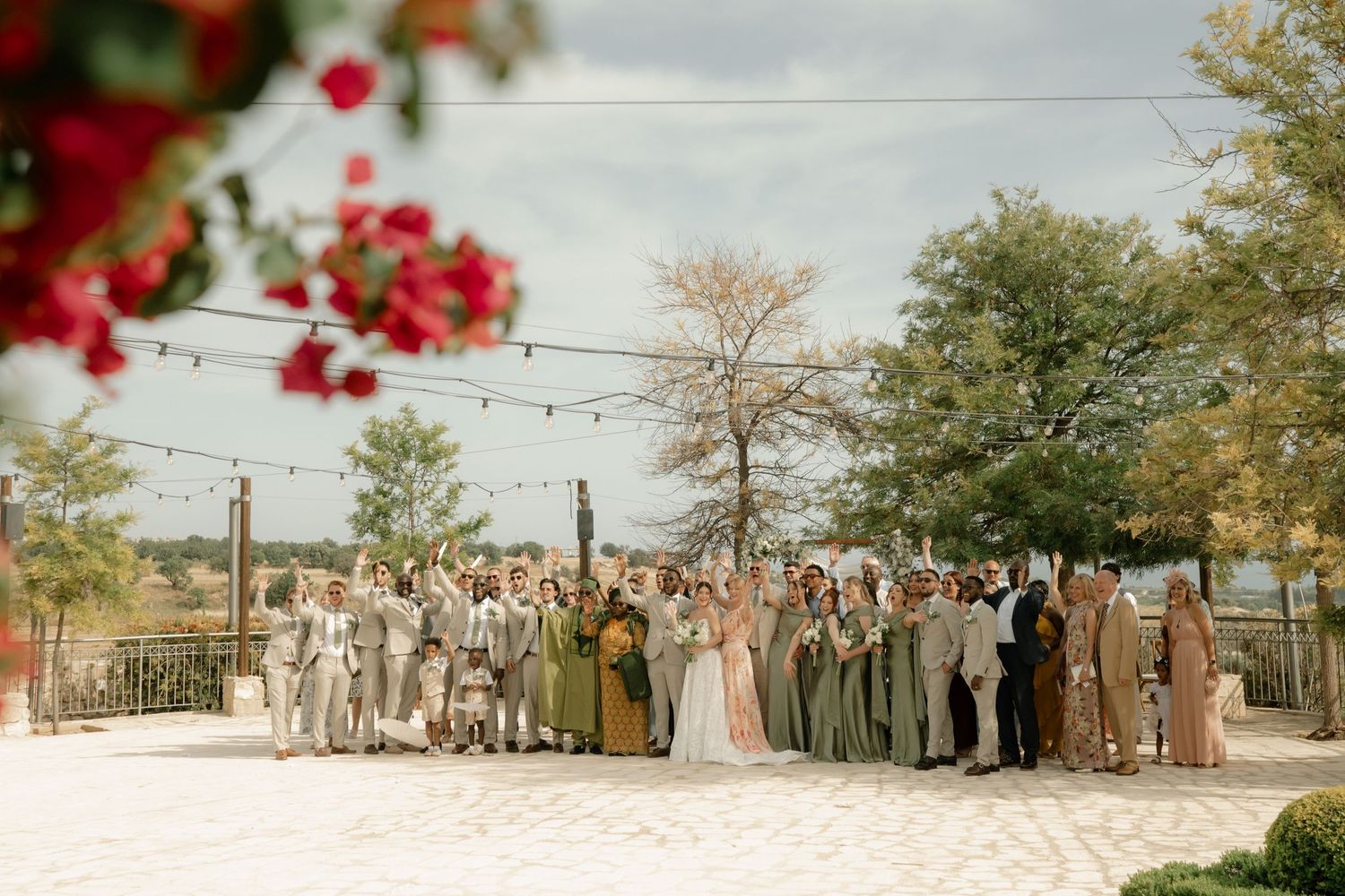 Large wedding party poses outdoors on white sand with string lights and trees at rustic venue.