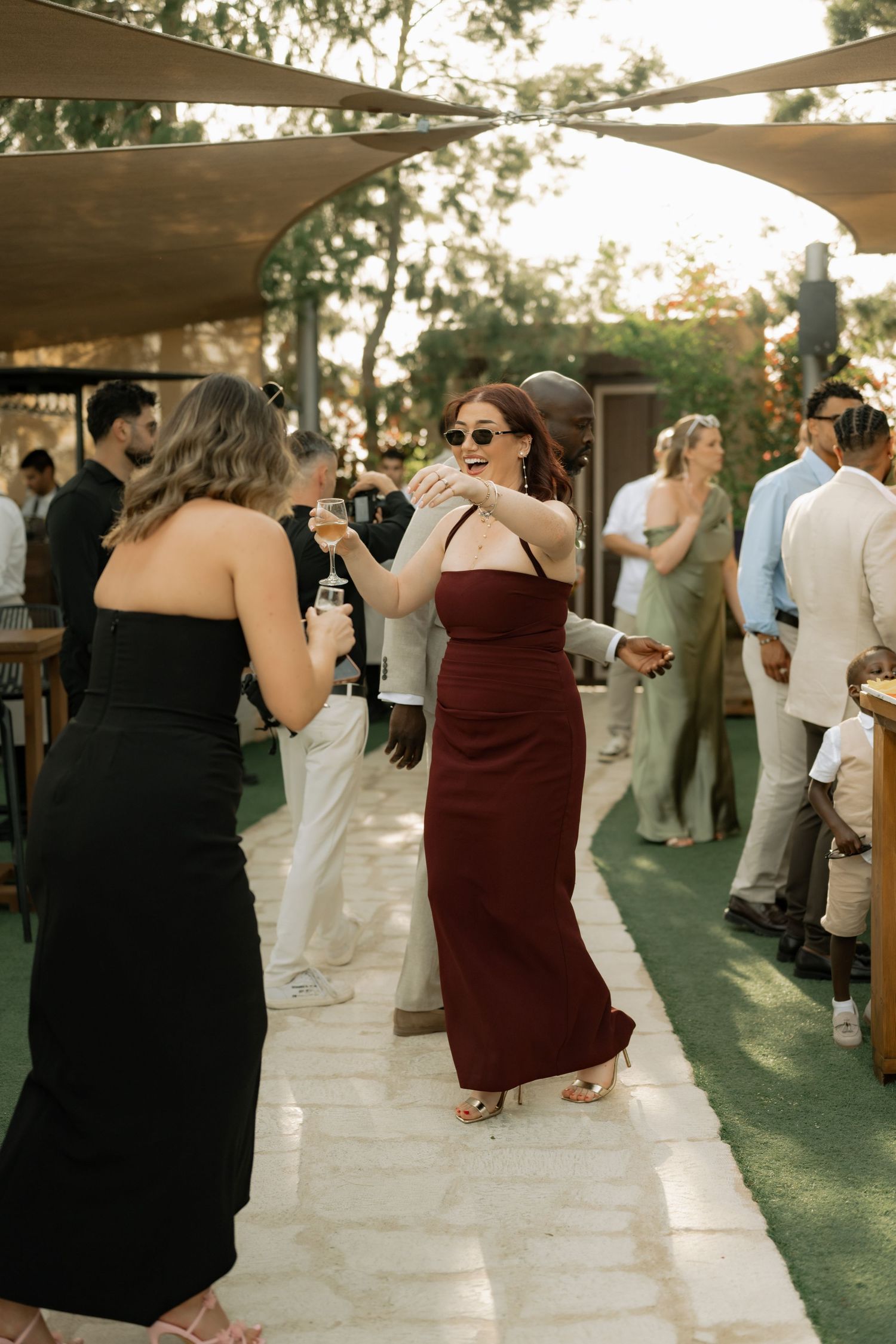 Two women in elegant dresses dancing at an outdoor wedding reception with guests mingling in the background.