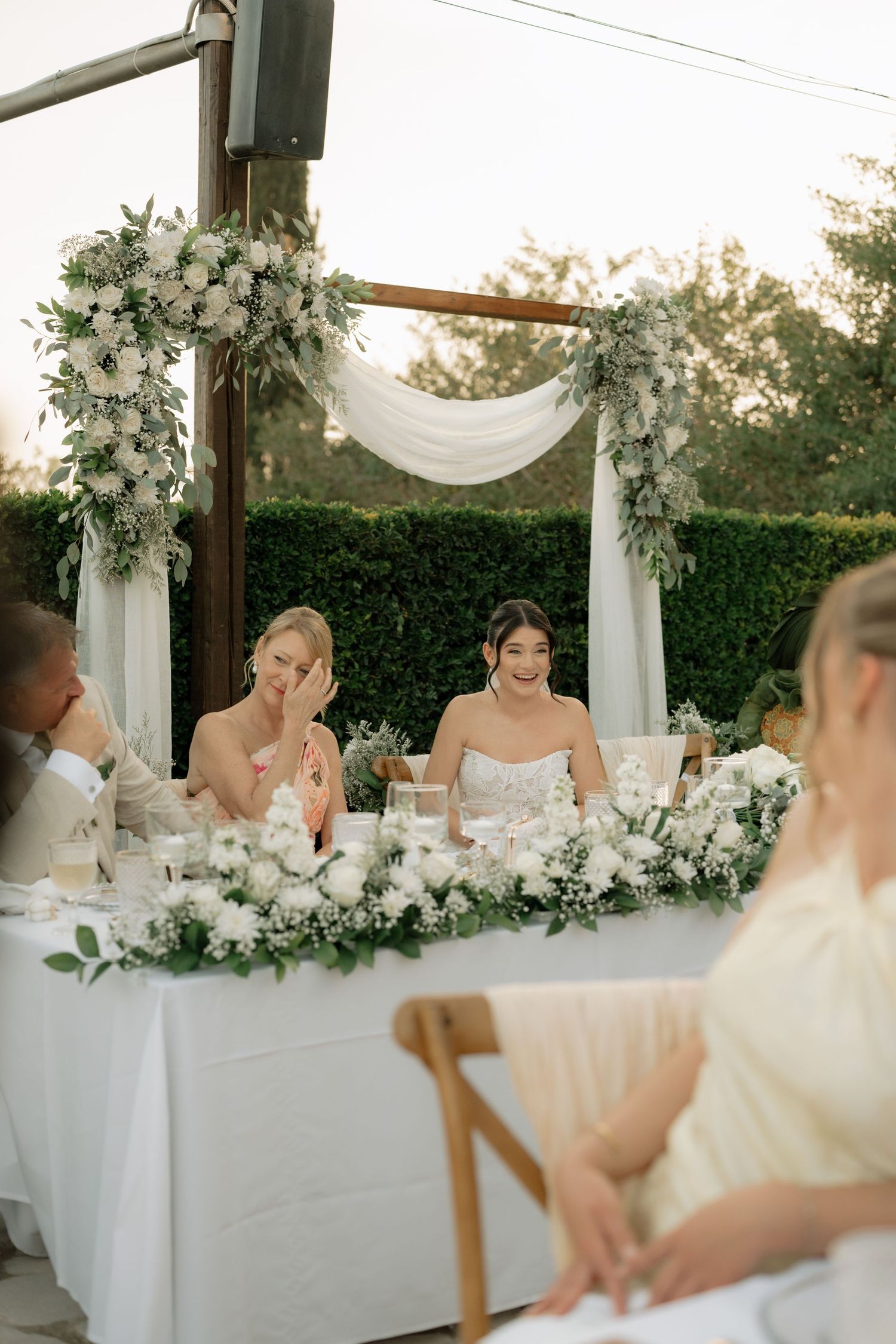 Bride and bridesmaid smiling at outdoor wedding reception table adorned with white floral arrangements and greenery.