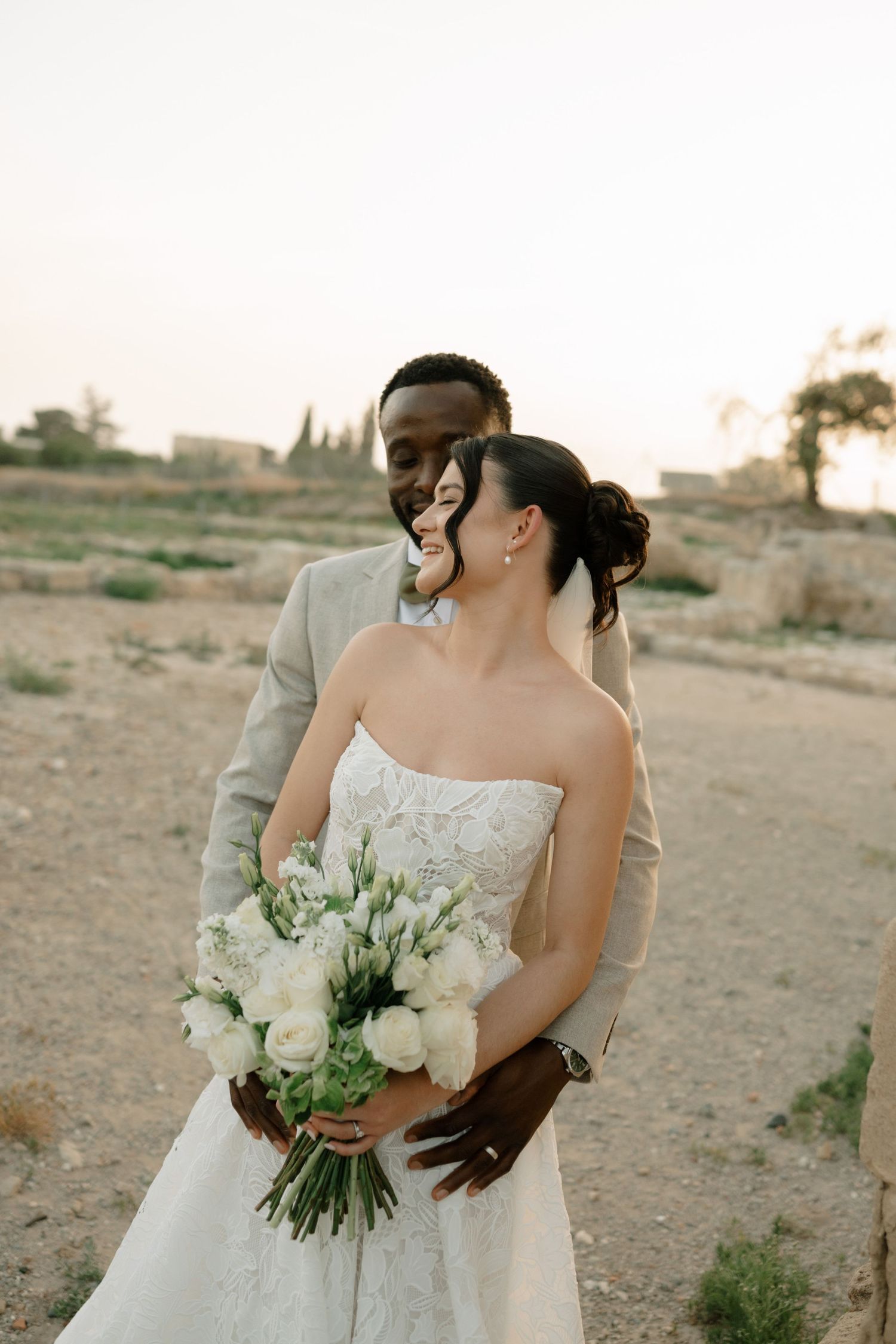 Interracial couple embracing at sunset, bride in strapless lace gown holding white bouquet, groom in beige suit.