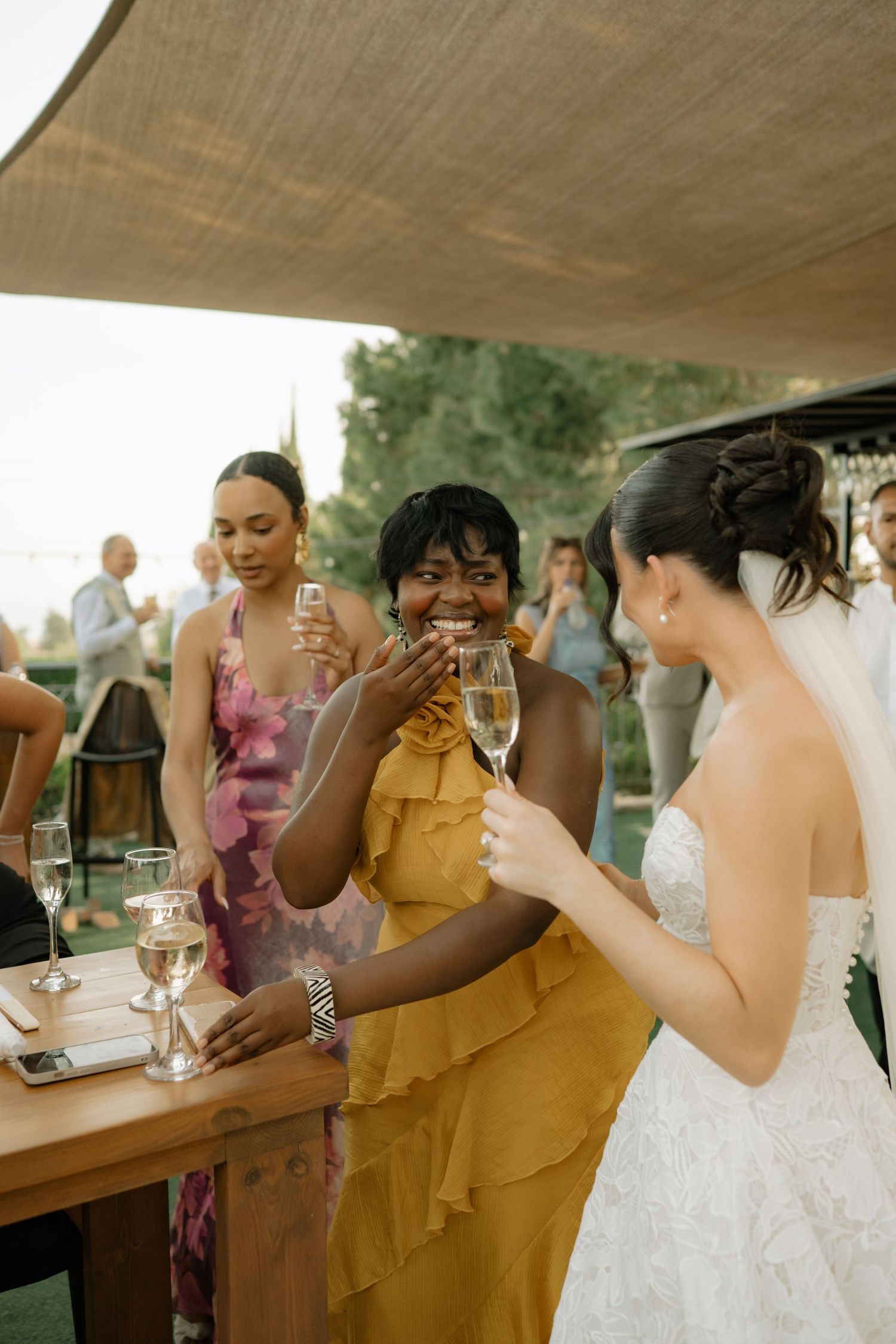 Bride in white lace gown toasting champagne with guest in yellow dress at outdoor wedding reception.