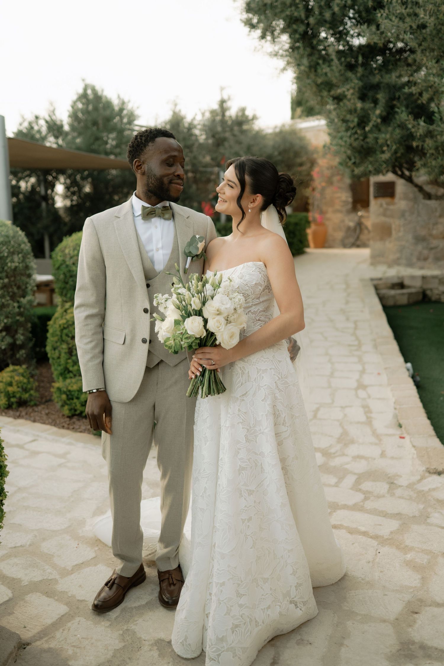 Interracial couple in wedding attire pose in garden, groom in beige suit, bride in white lace gown holding bouquet.