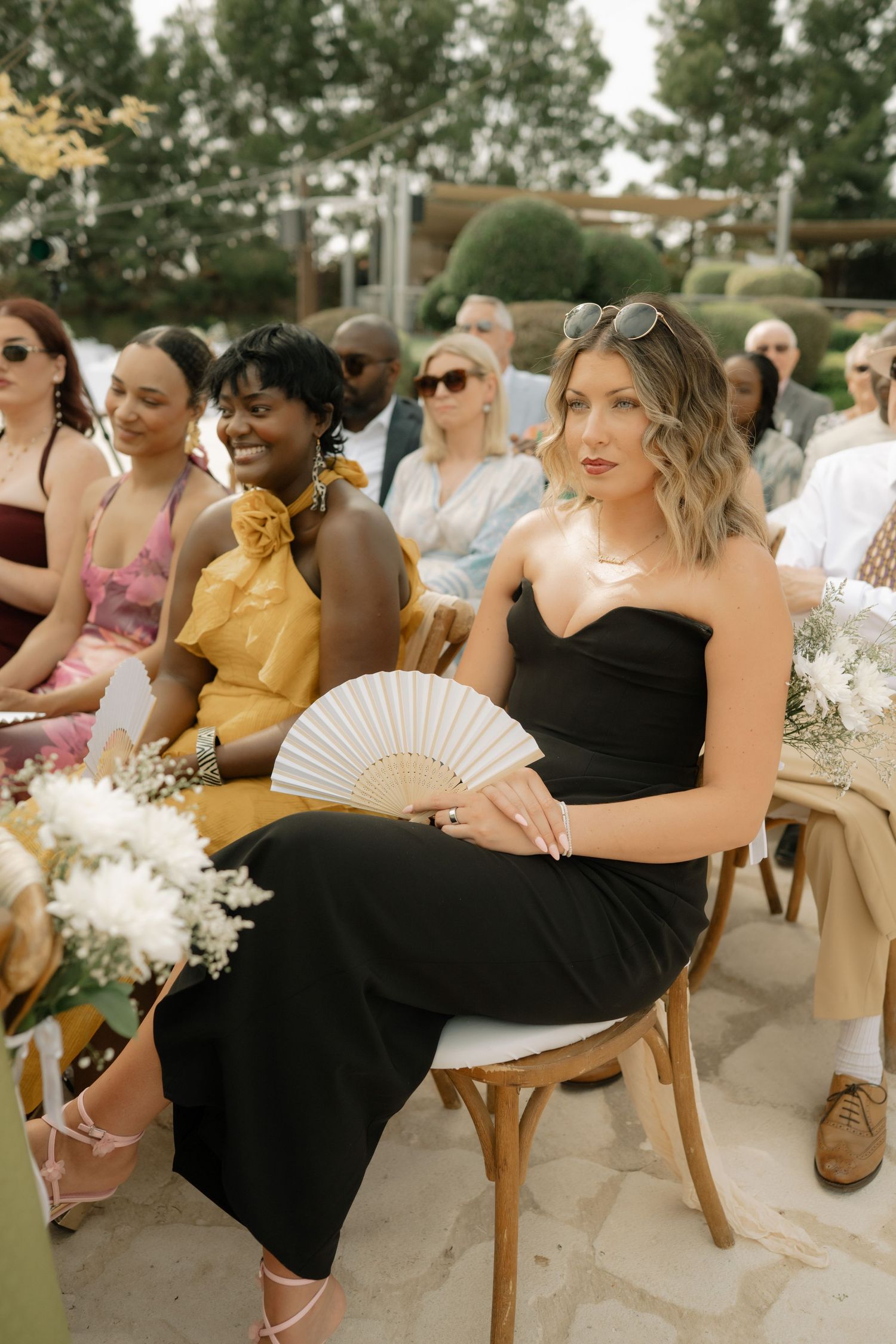 Guests seated outdoors at a wedding ceremony, woman in black strapless dress holding a white hand fan.