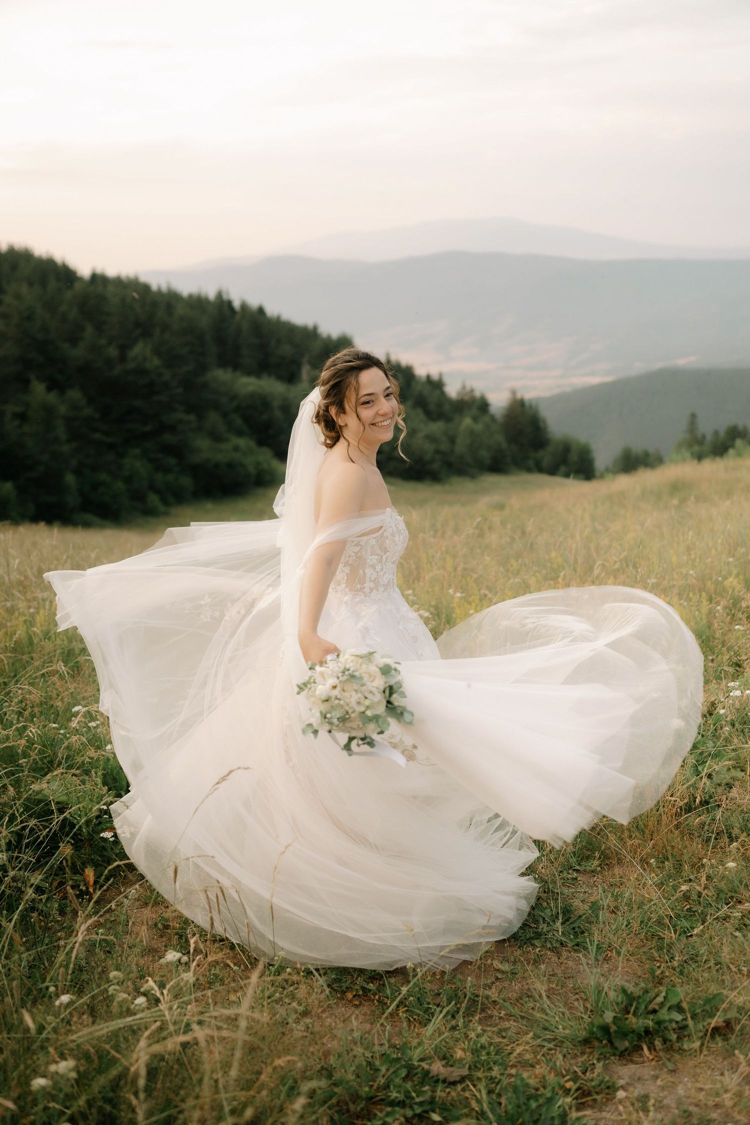 A flowing white wedding dress twirls gracefully in a mountain meadow with rolling hills and forests in the background.