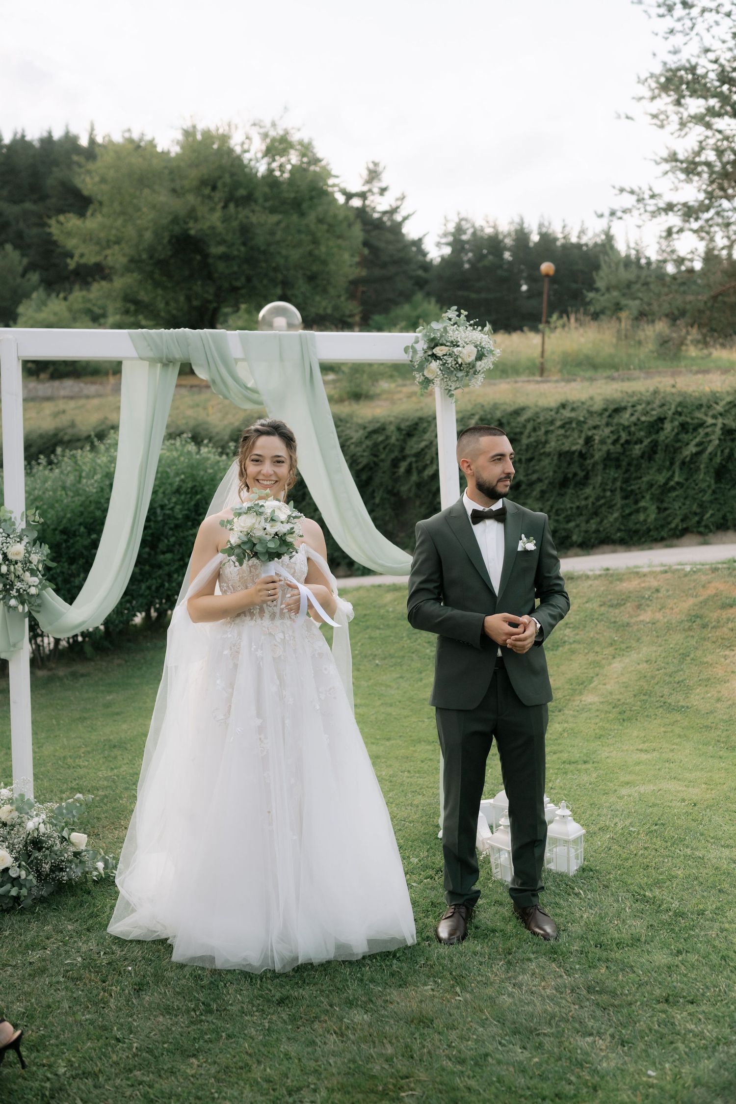 A romantic outdoor wedding scene with white draped fabric decor and floral arrangements on a grassy lawn.