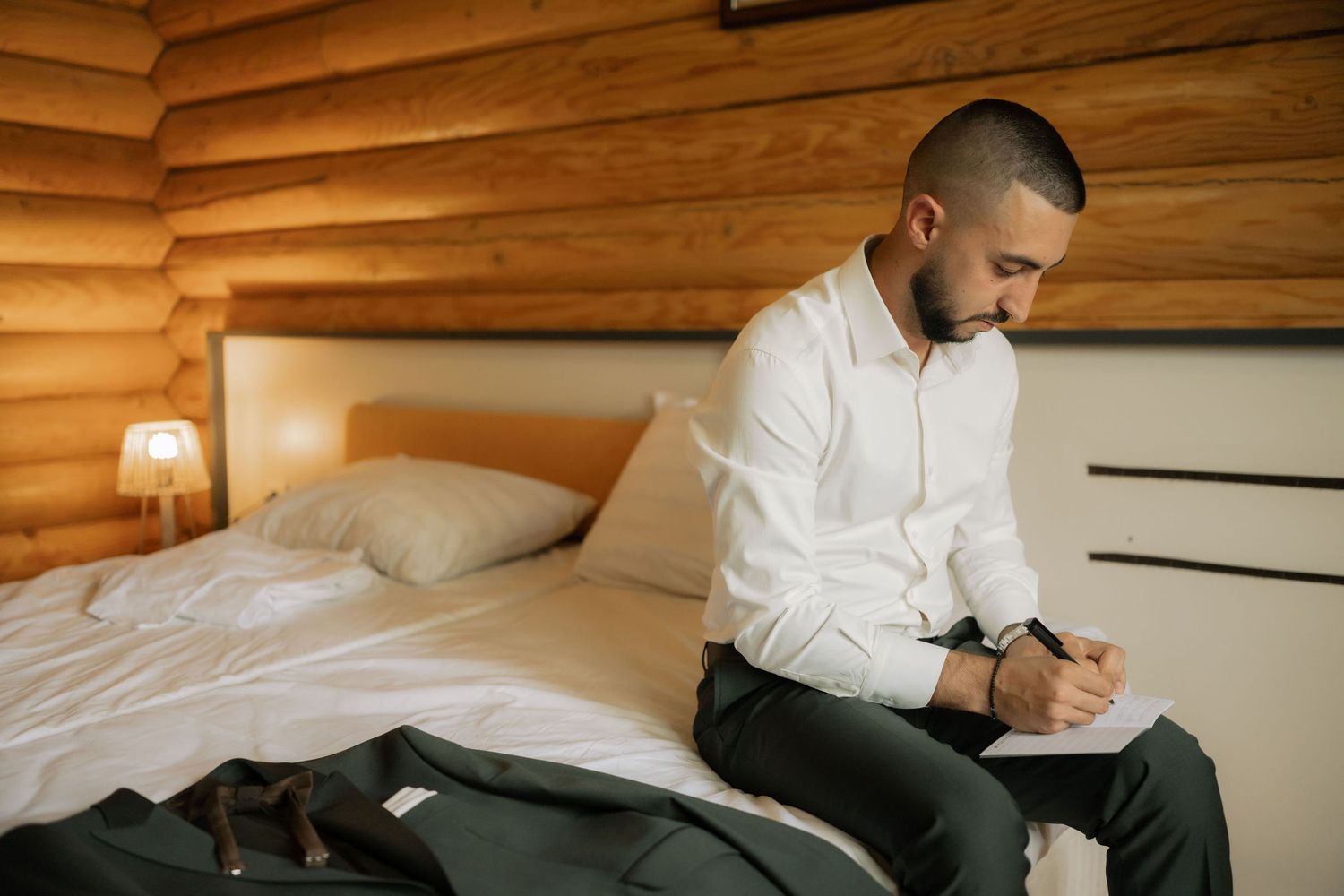 Person in white shirt and black pants sits on bed in rustic log cabin bedroom writing in notebook.